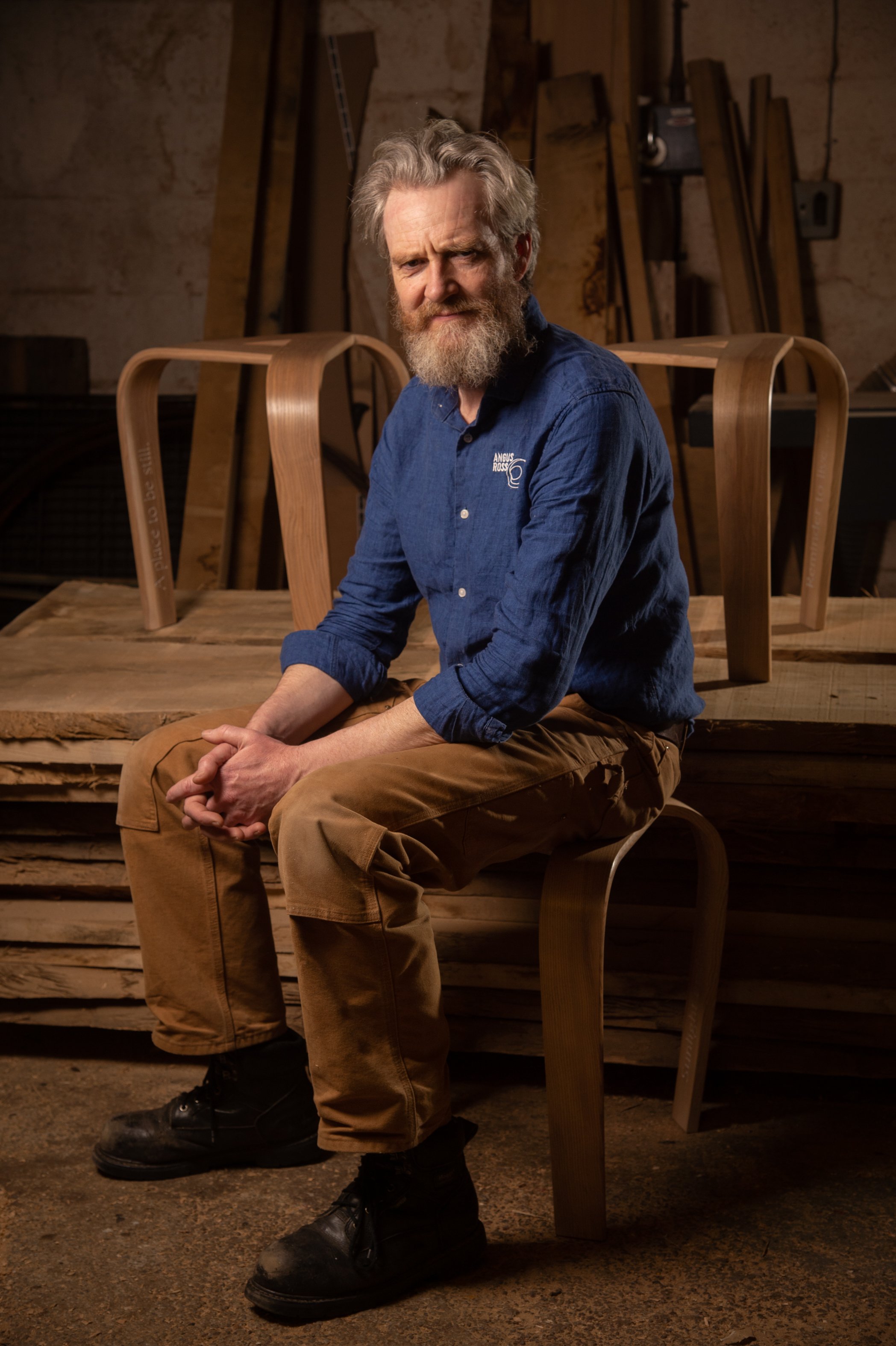 Angus Ross wearing a blue shirt and tan pants, sitting on a wooden stool in a woodworking shop. There are bentwood chair parts on the table behind him.