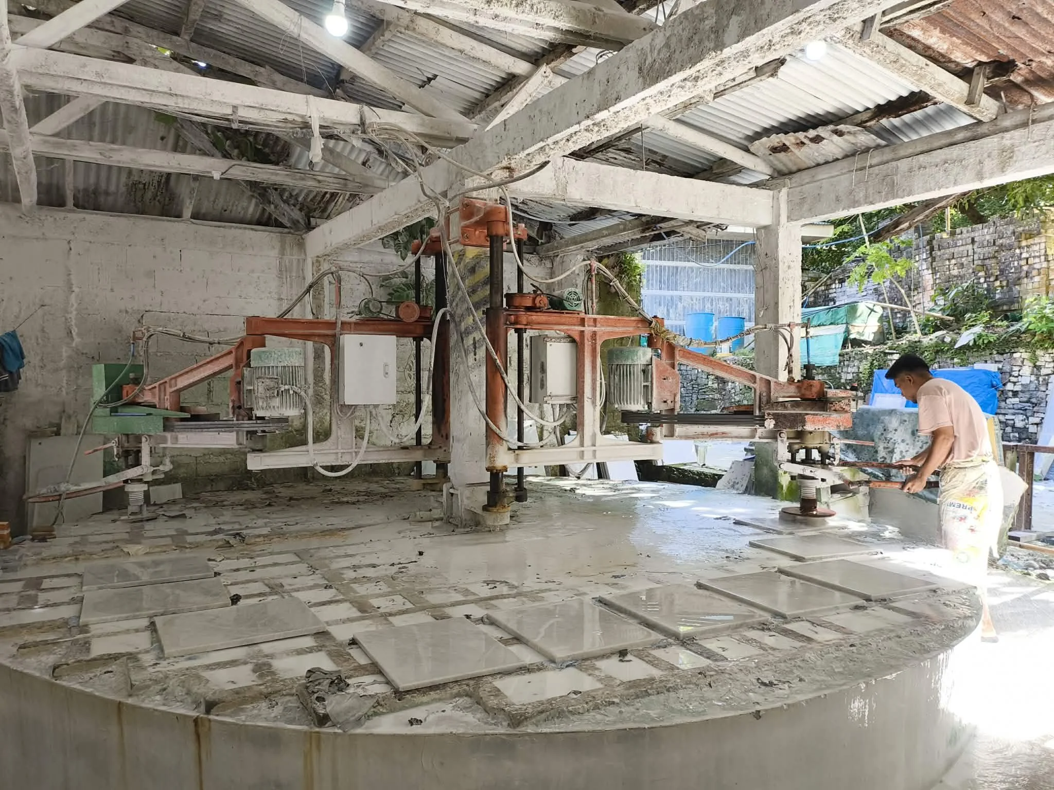 A worker on the right is polishing or cleaning a large stone table using a polishing machine. The table has multiple rectangular stone slabs arranged on it. The setting appears to be a workshop with unfinished, rustic surroundings and a corrugated metal roof.