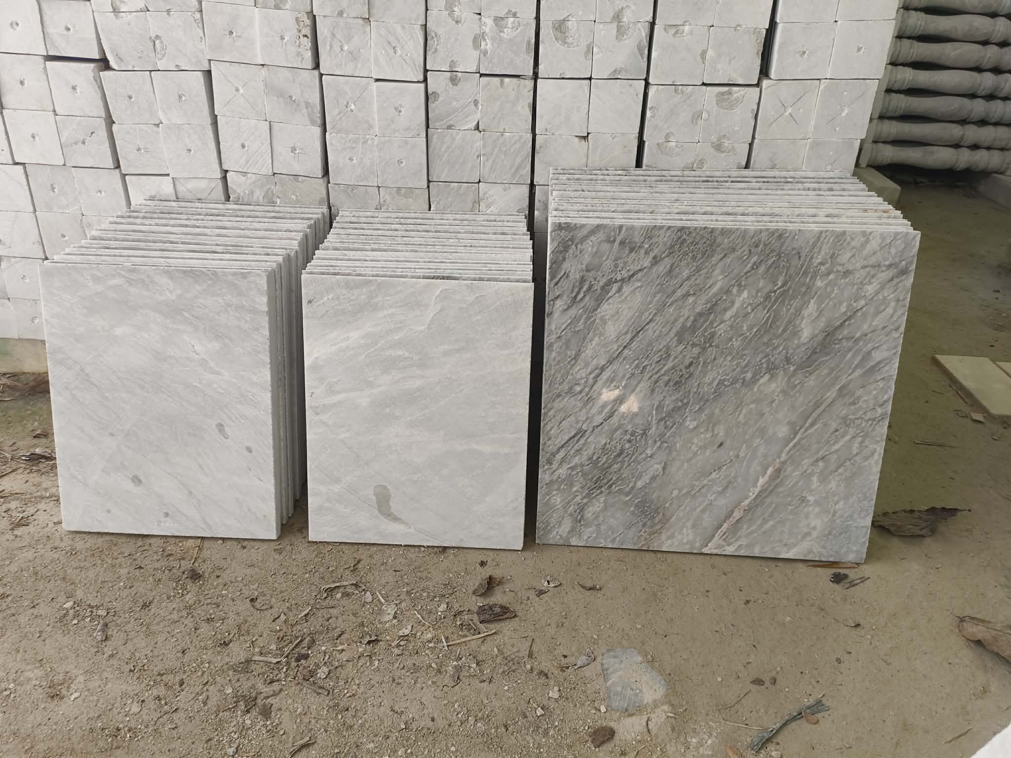 Stacks of marble tiles in different shades of gray on a dirt ground, with some stored marble blocks in the background.