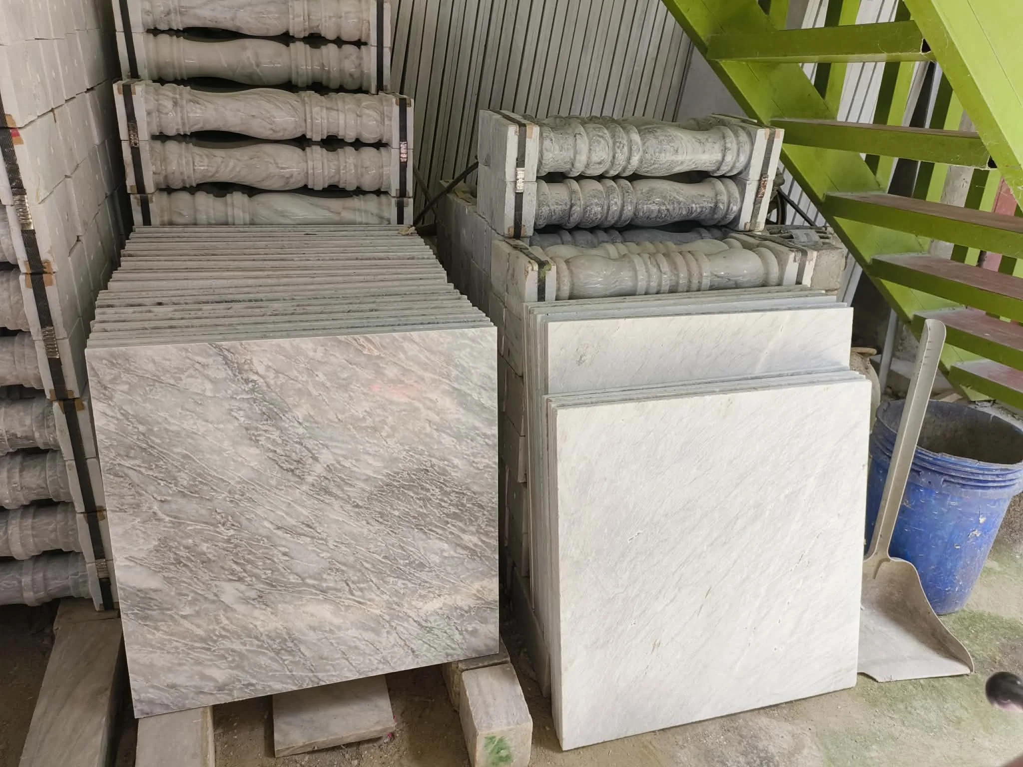 Stacks of marble tiles and marble balusters in a storage area, with a blue bucket and a shovel nearby.