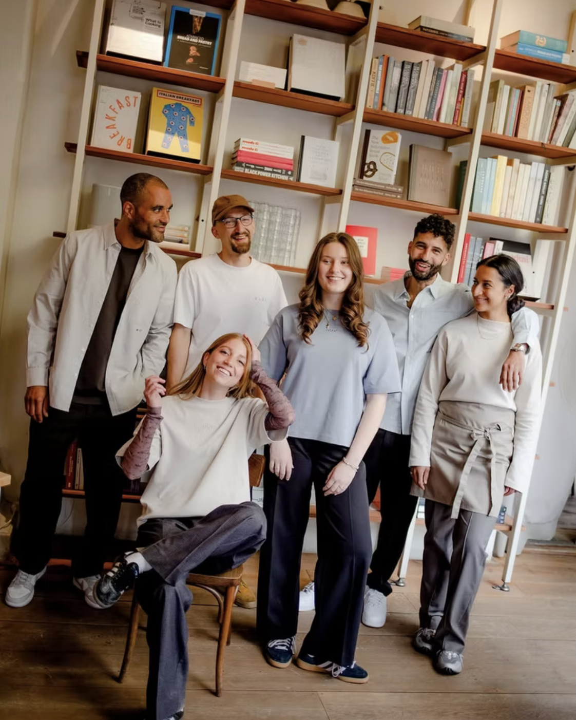 A diverse group of seven young adults smiling and posing indoors in front of a bookshelf filled with books.