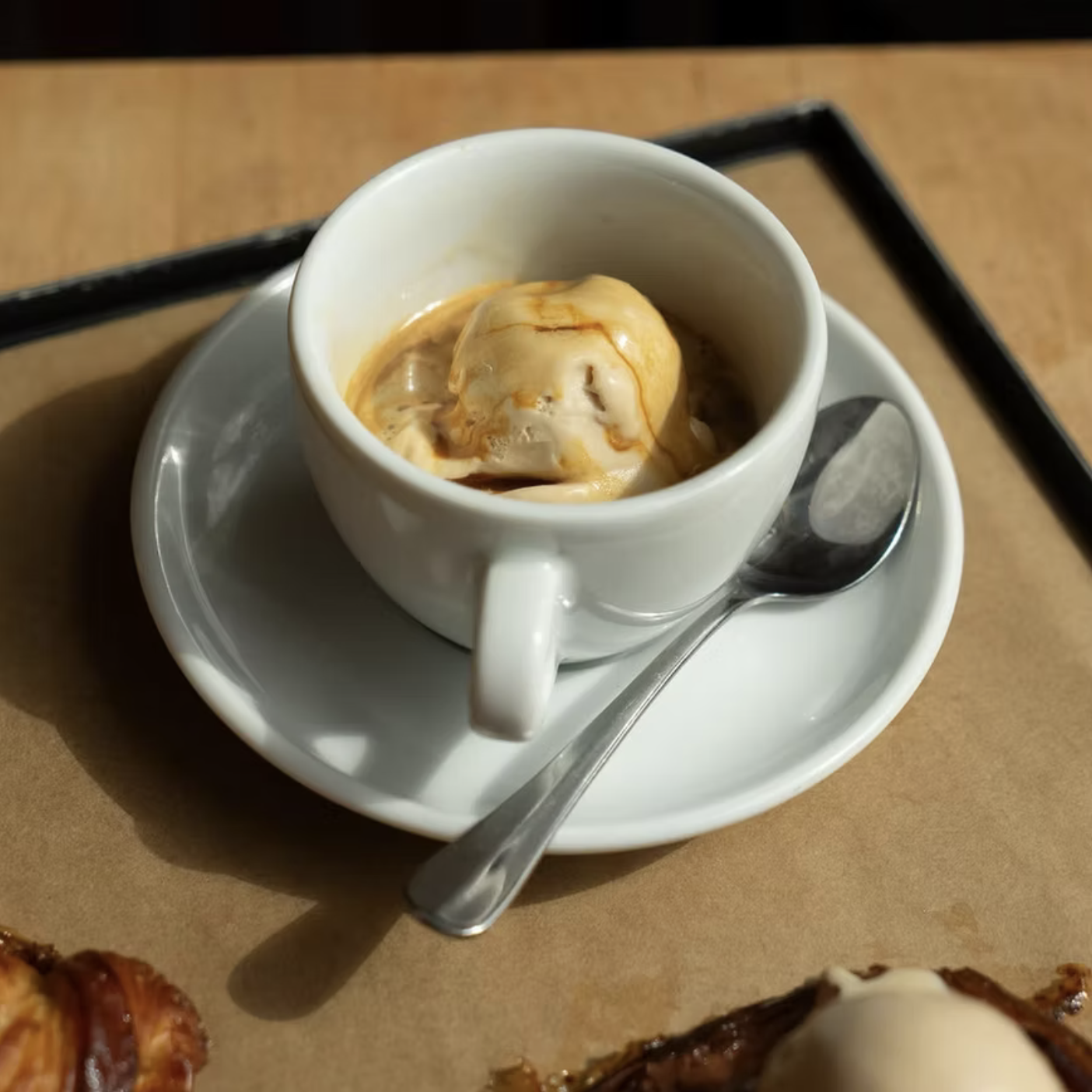 A small white cup of vanilla ice cream topped with caramel sauce on a saucer with a metal spoon, on a wooden table.
