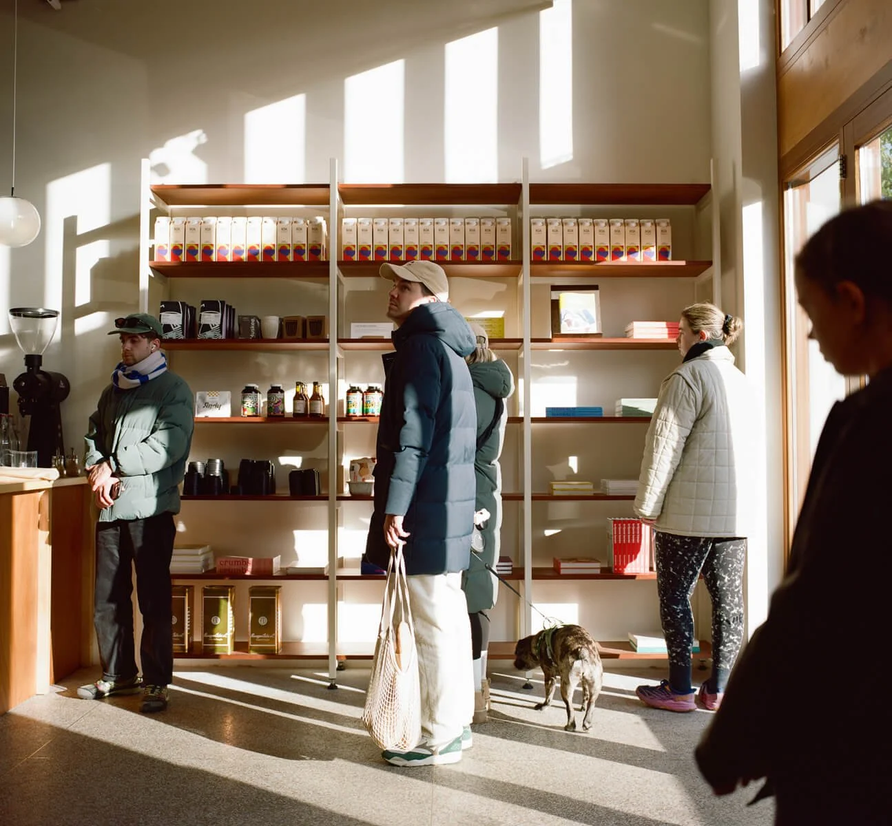 People ordering at a coffee shop counter with sunlight streaming through large windows, shelves with products behind them, and a person holding a dog.