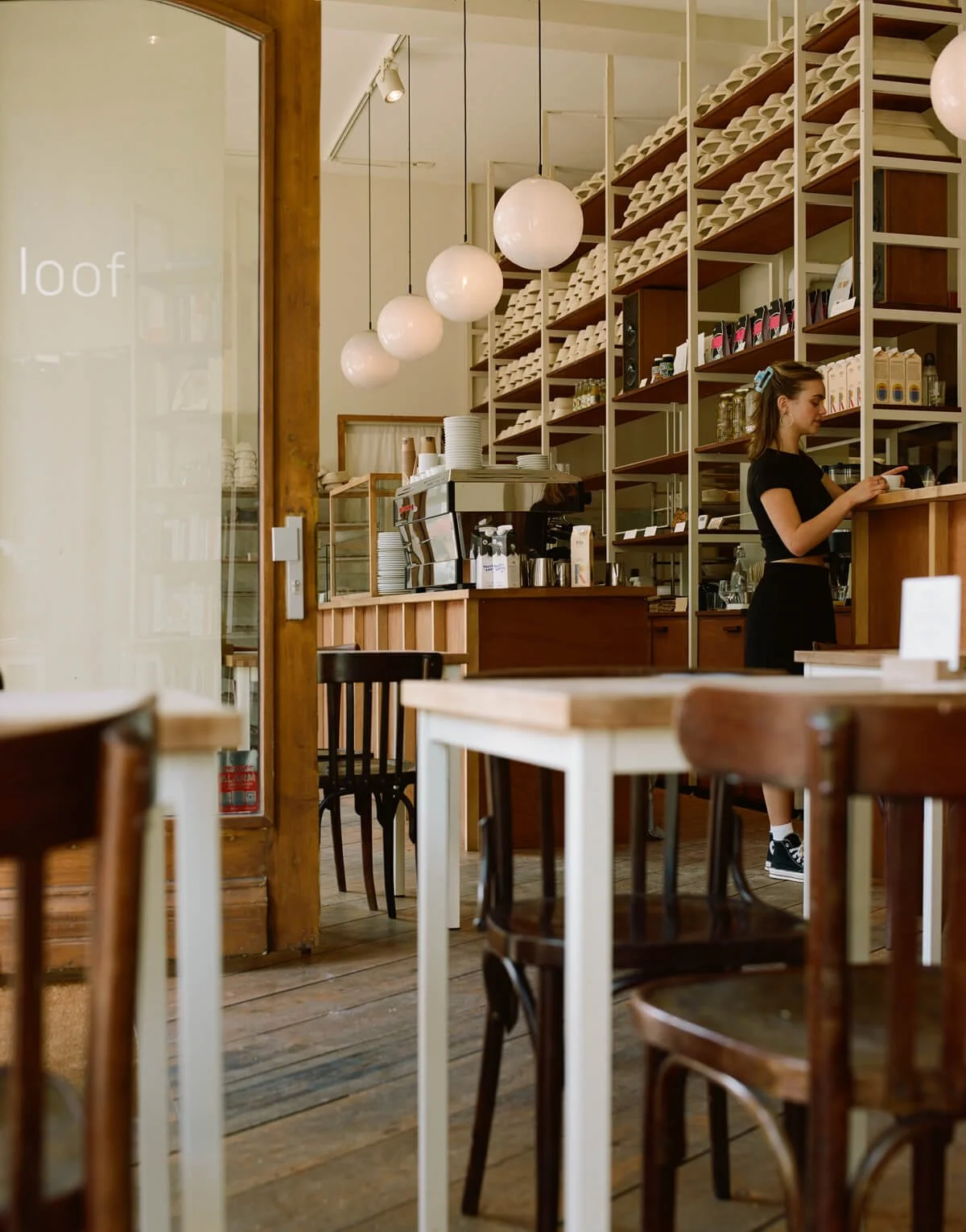 Interior of a cozy coffee shop with wooden tables and chairs, a barista behind the counter, white spherical pendant lights, and shelves with coffee supplies.