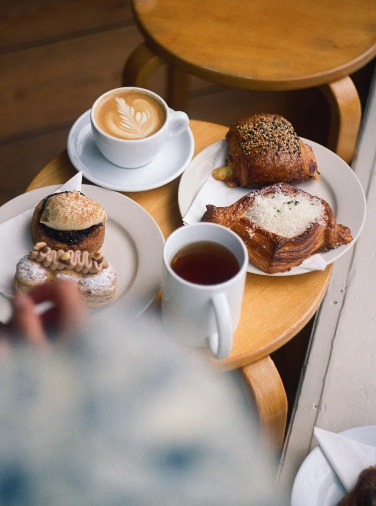 A wooden table with two cups of coffee, one latte art, a mug of tea, and a white plate with pastries, including a croissant and a square pastry with sesame seeds.