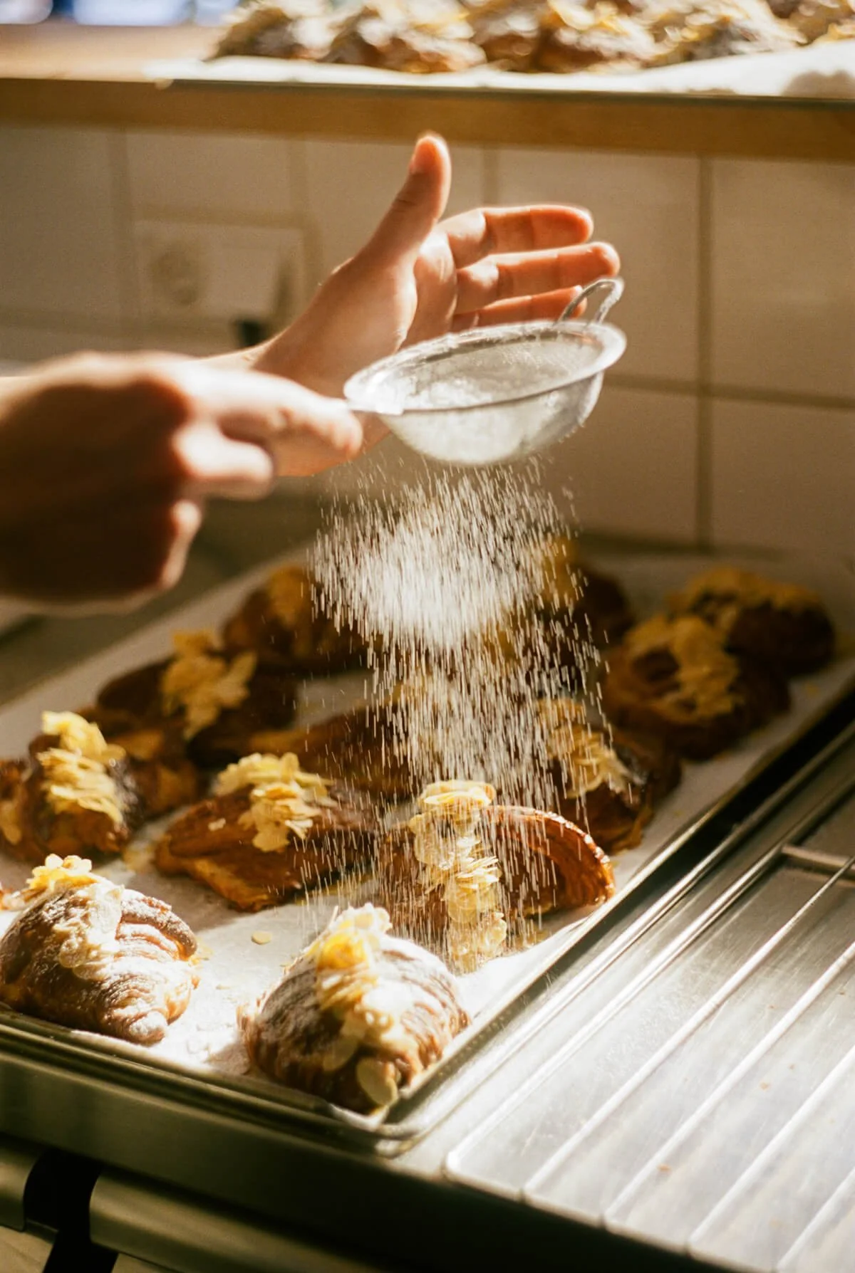 A person sifting powdered sugar over freshly baked cookies decorated with almond slices on a baking sheet.