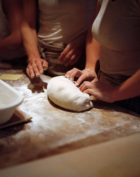 People working together to knead and shape pizza dough on a floured wooden surface.