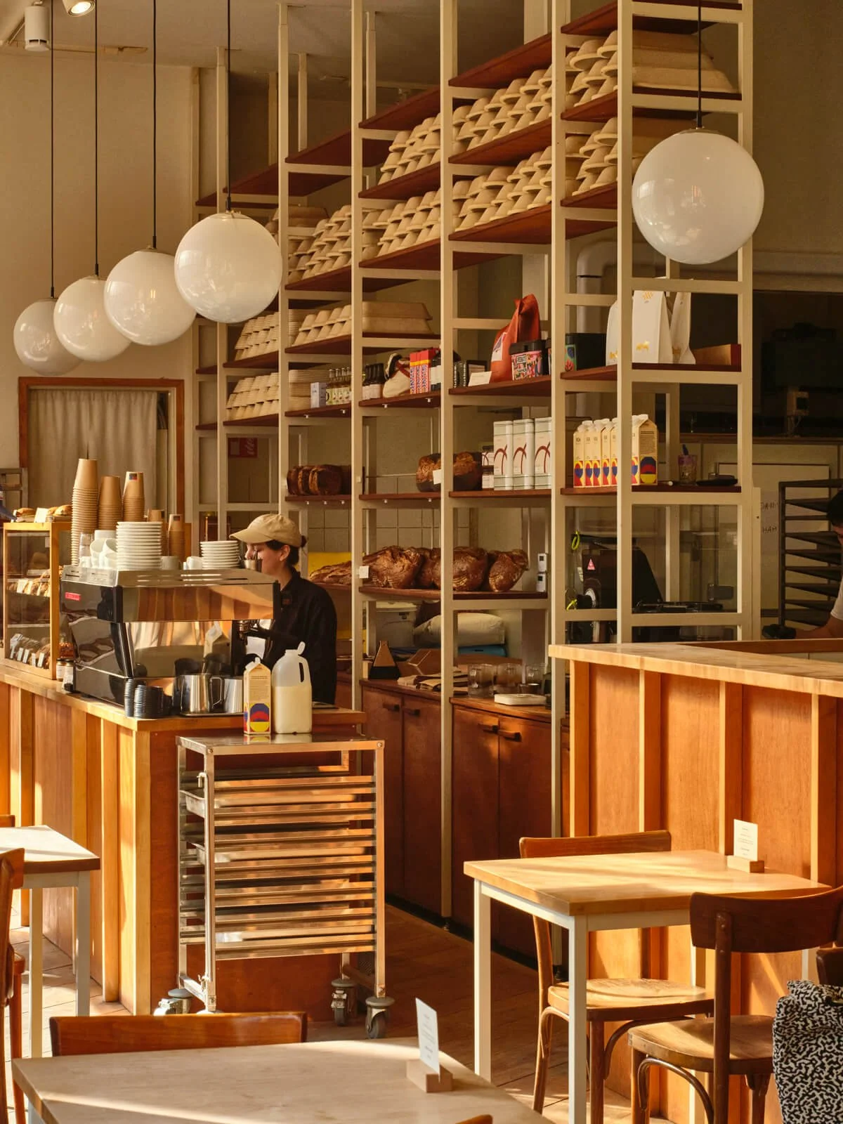 Interior of a bakery or cafe with wooden furniture, a barista behind the counter, shelves with baked goods, white globe pendant lights, and tables for customers.