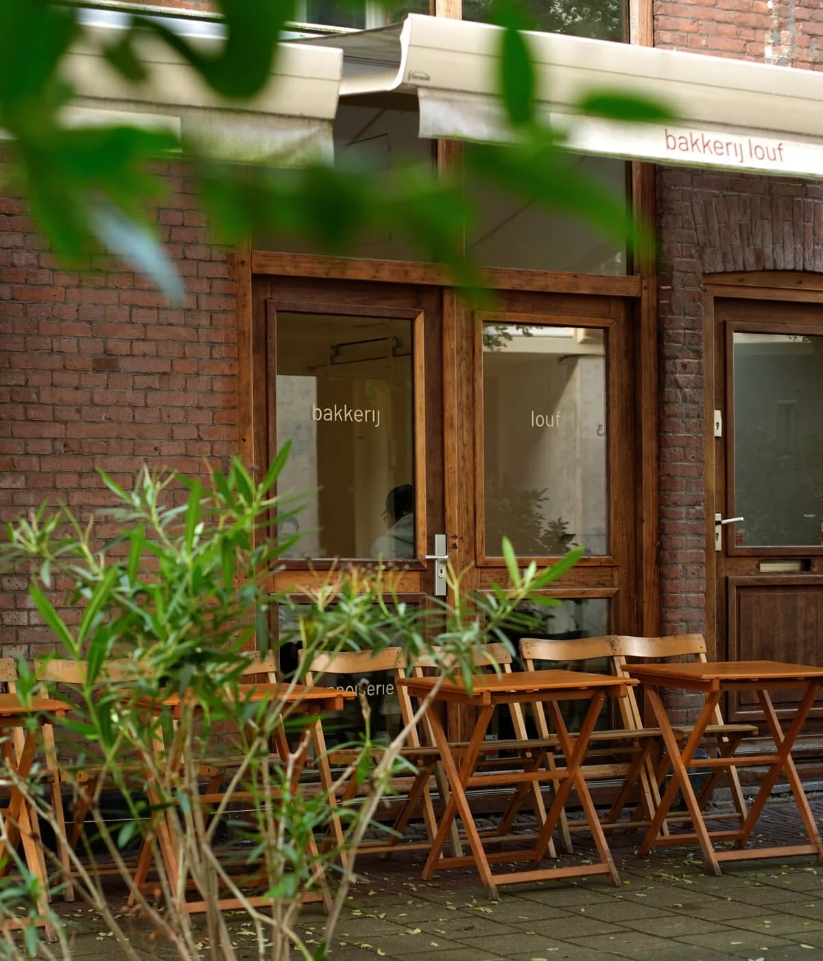 Exterior view of a bakery and lunch shop with wooden framed windows and a door, outdoor seating with wooden chairs and tables, brick building, green plants in foreground, awning above with partial text "bakkerij" and "lunch" visible.