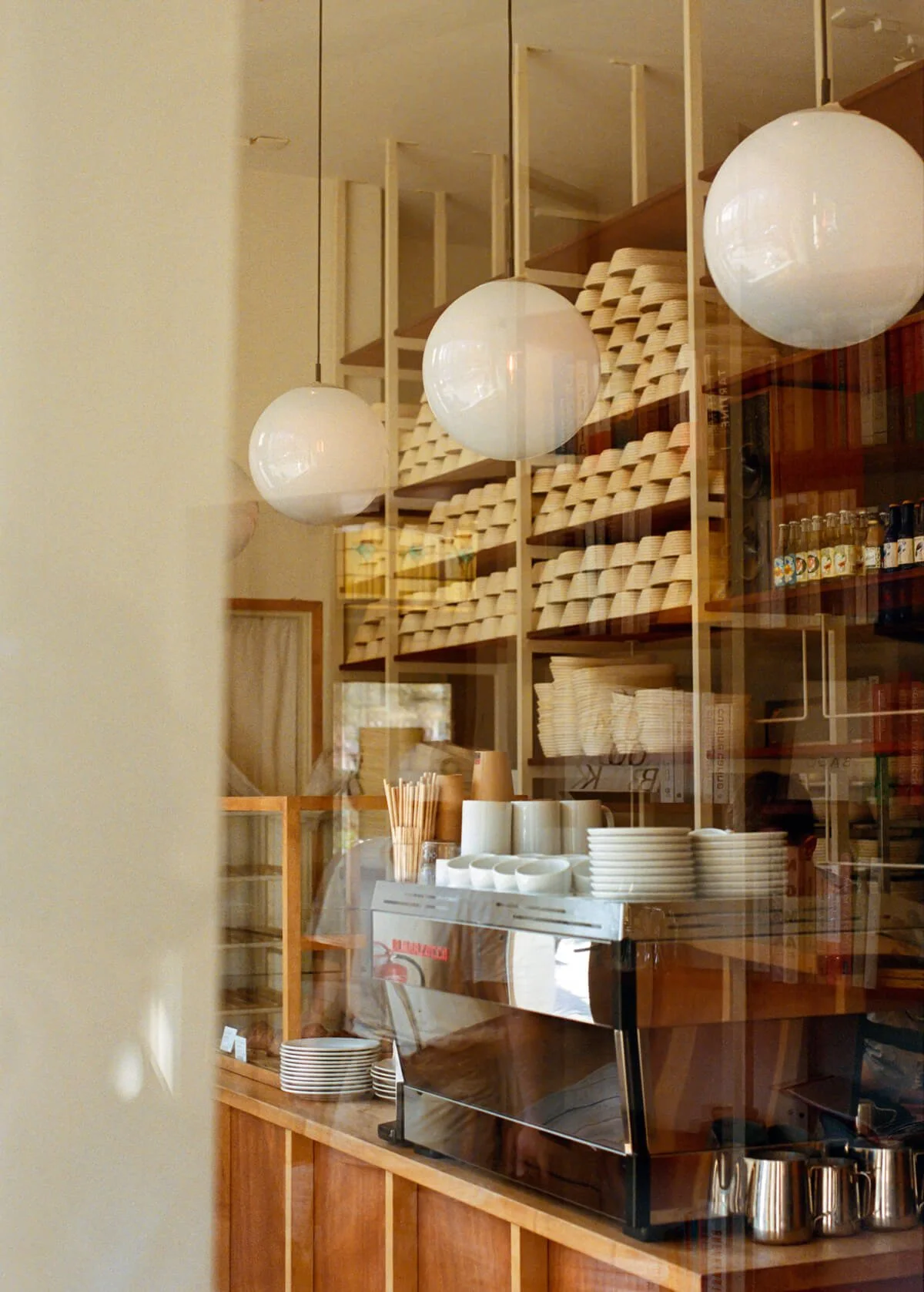 Inside a cafe, with hanging spherical white lights, wooden shelves filled with dishes, cups and containers, and a stainless steel espresso machine on a wooden counter.