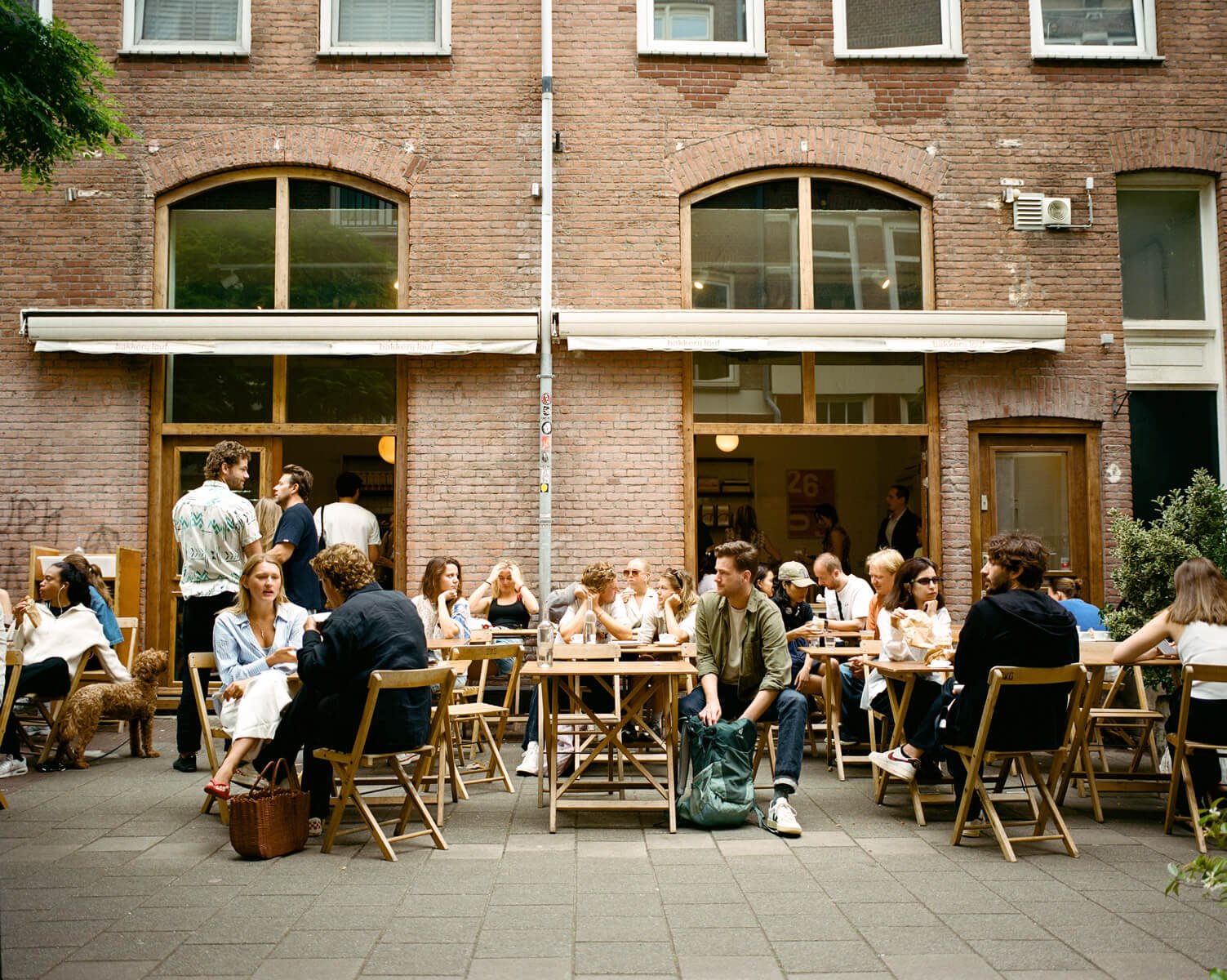 People sitting at outdoor tables in front of a cafe or restaurant on a city street, with brick buildings in the background.