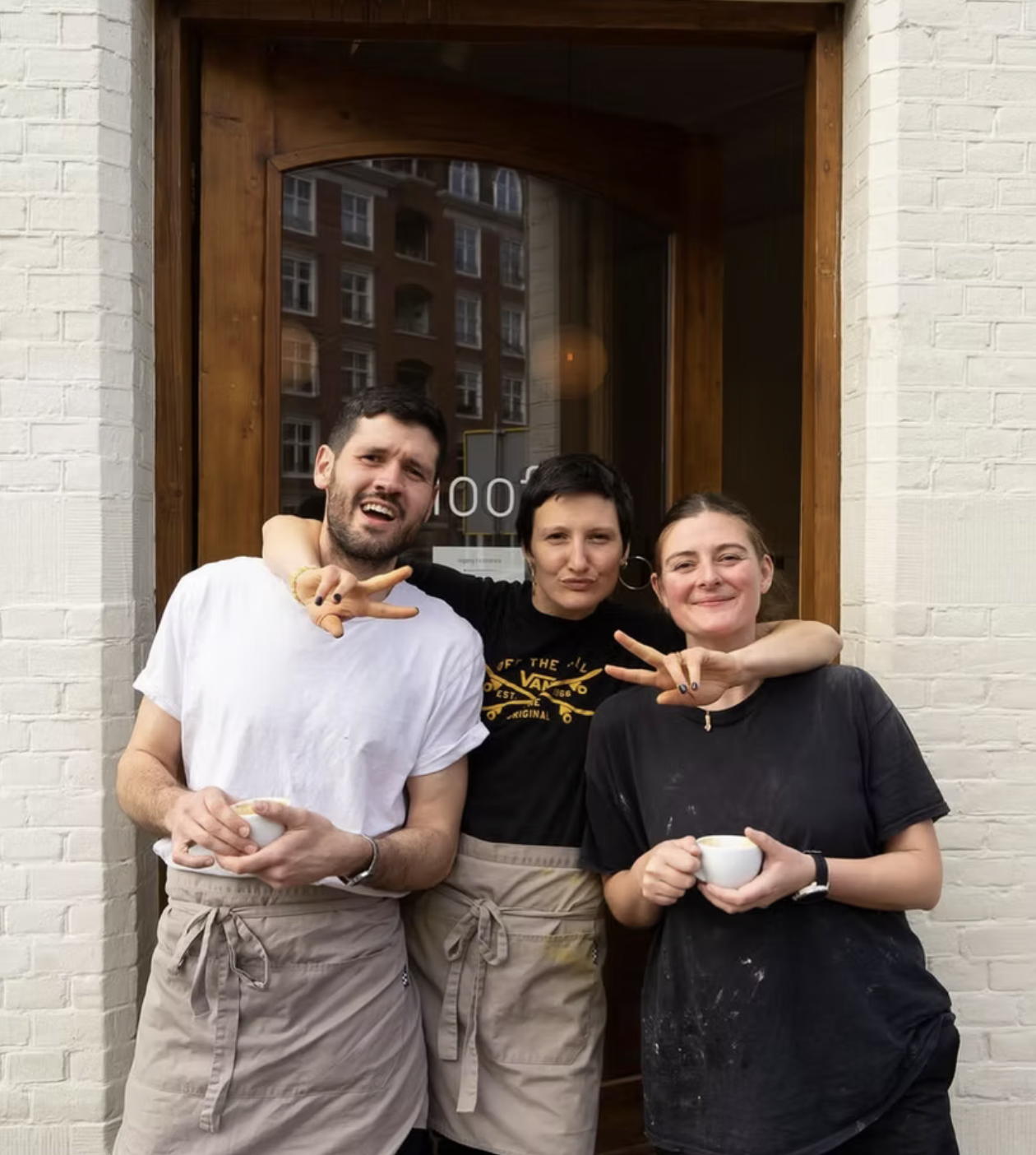 Three people standing outside a restaurant, smiling and making peace signs, holding coffee cups. The group includes a man with a white t-shirt, a woman in a black t-shirt, and a woman in a black smock, in front of a brick wall with a wooden door and 