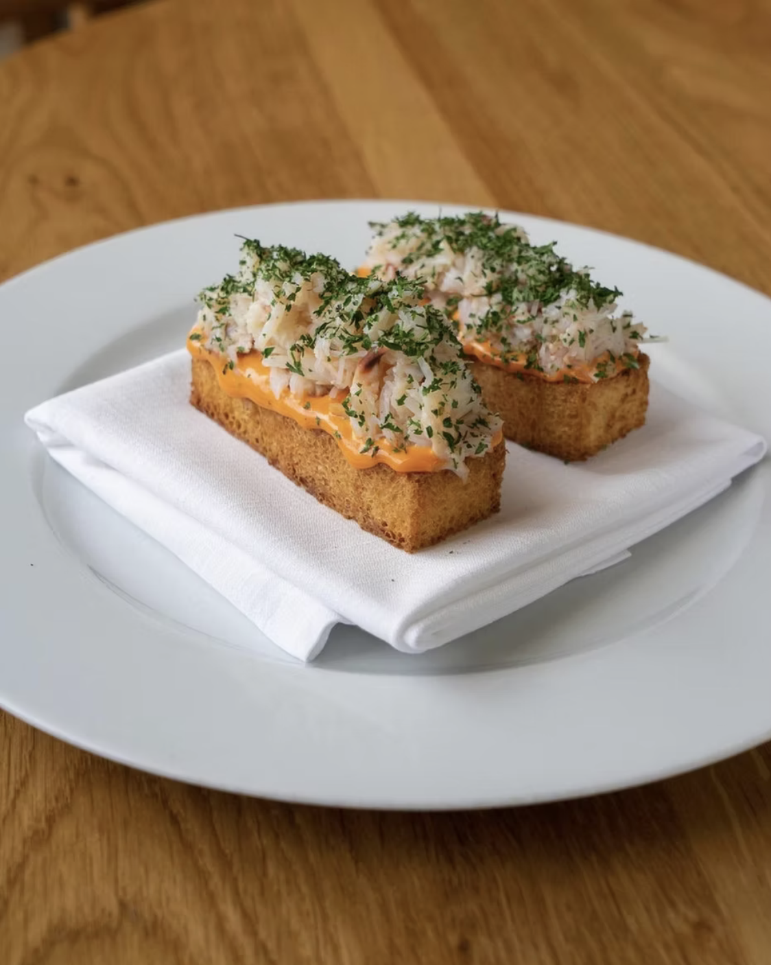 Two pieces of toast topped with a spread of orange sauce, shredded crab meat, chopped herbs, served on a white plate with a napkin.