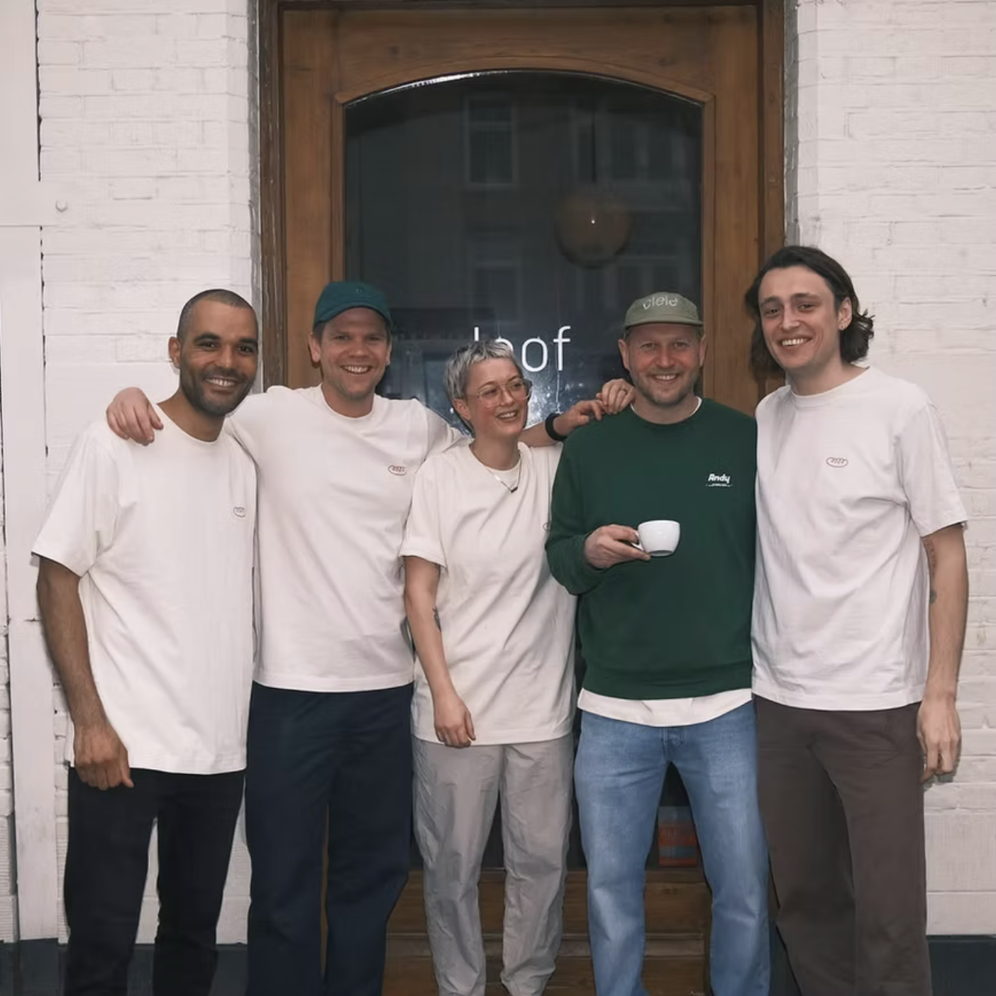 Group of five people, smiling and standing together in front of a wooden door with a glass window, in an indoor setting with white brick walls. The person in the center is holding a coffee cup.