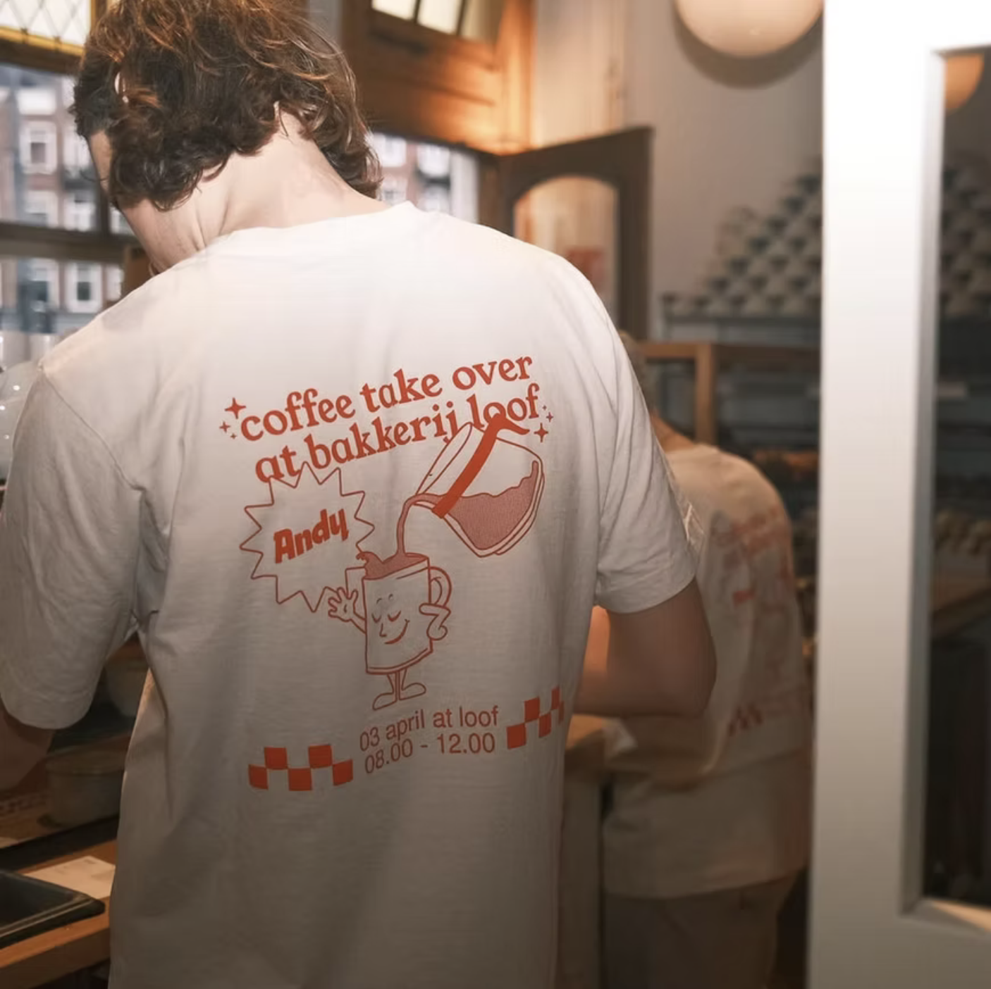 Back of a person wearing a white t-shirt with a graphic of a coffee mug pouring coffee, event details, and the text 'coffee take over at bakkerij loof Andy'.
