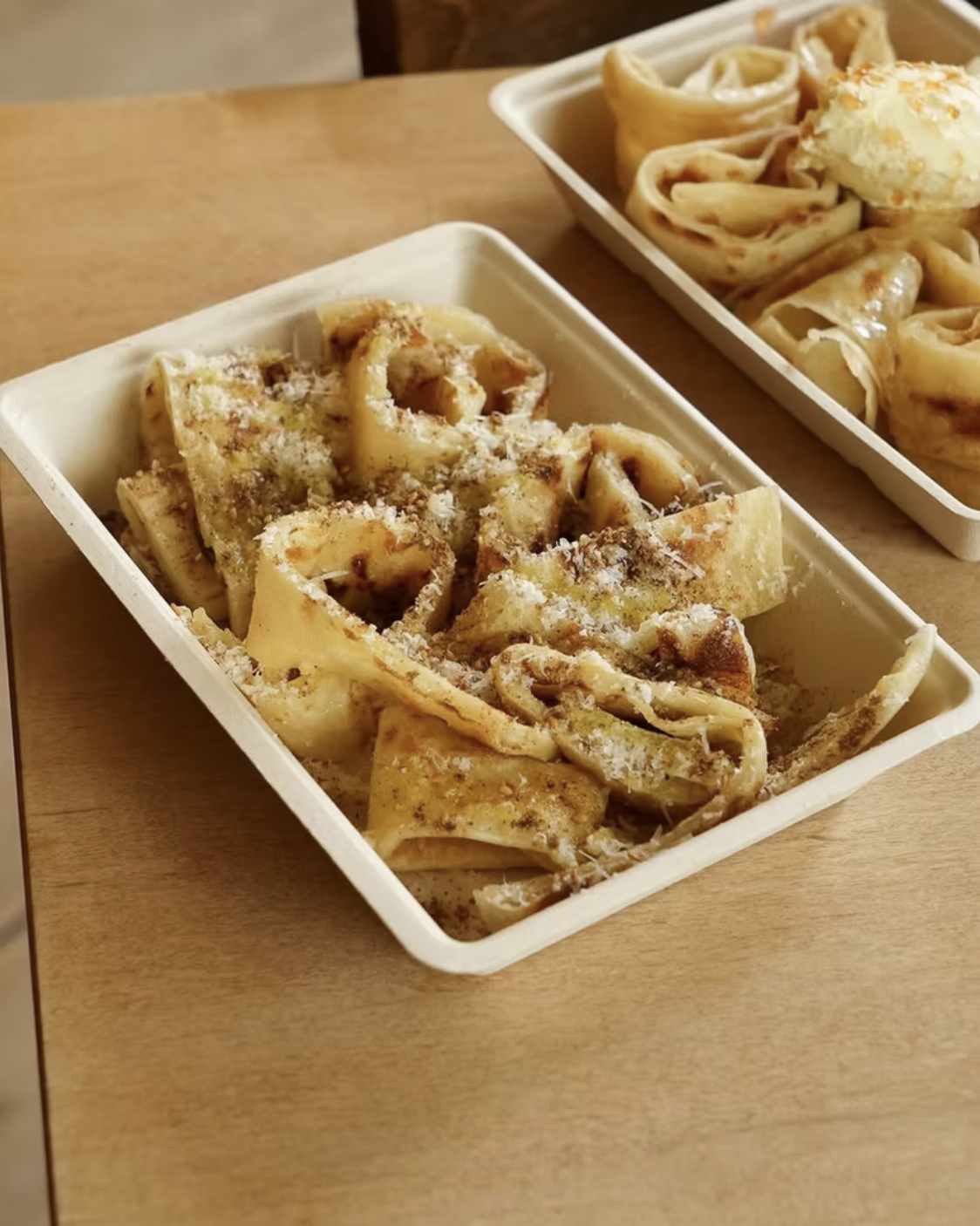 Two white rectangular dishes with cooked pasta topped with grated cheese on a wooden table.