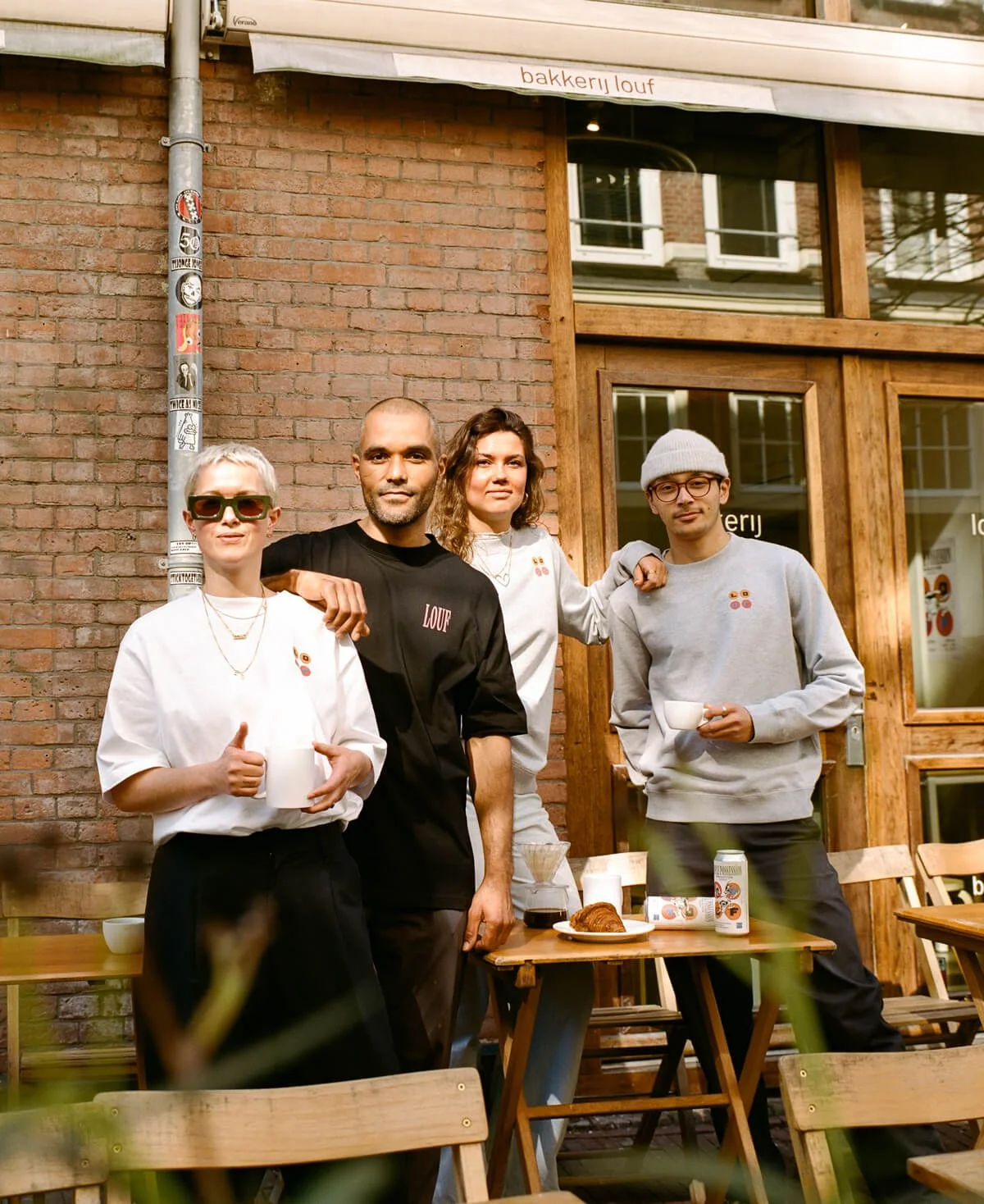 Four people standing outside a coffee shop, smiling, and holding cups, with a table and food items in front of them.