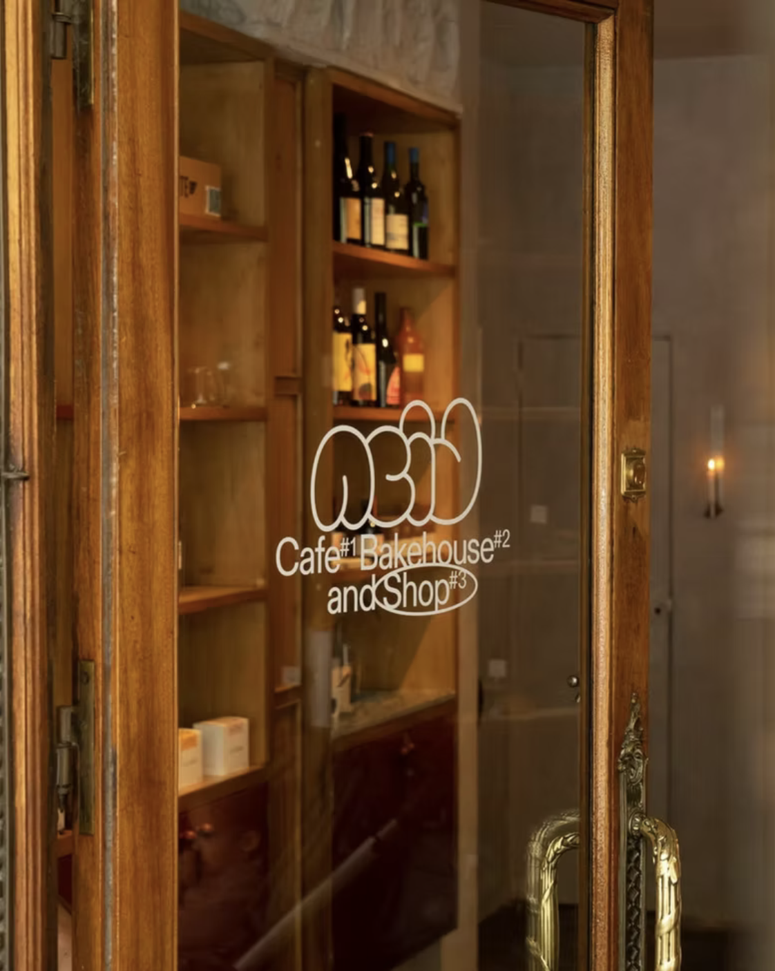 Glass door with wooden frame and golden handle, has a sign that reads 'AOSA Cafe #1 Bakehouse #2 and Shop #3', showing inside shelves with wine bottles and other items.
