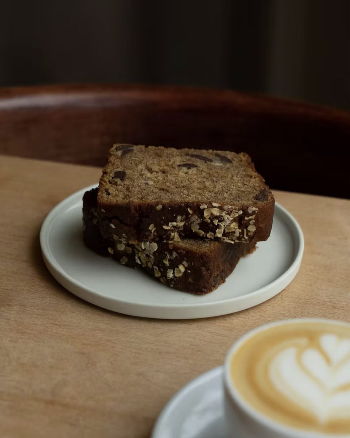 A slice of banana bread with nuts on a small white plate, with a cup of coffee with latte art in the foreground.