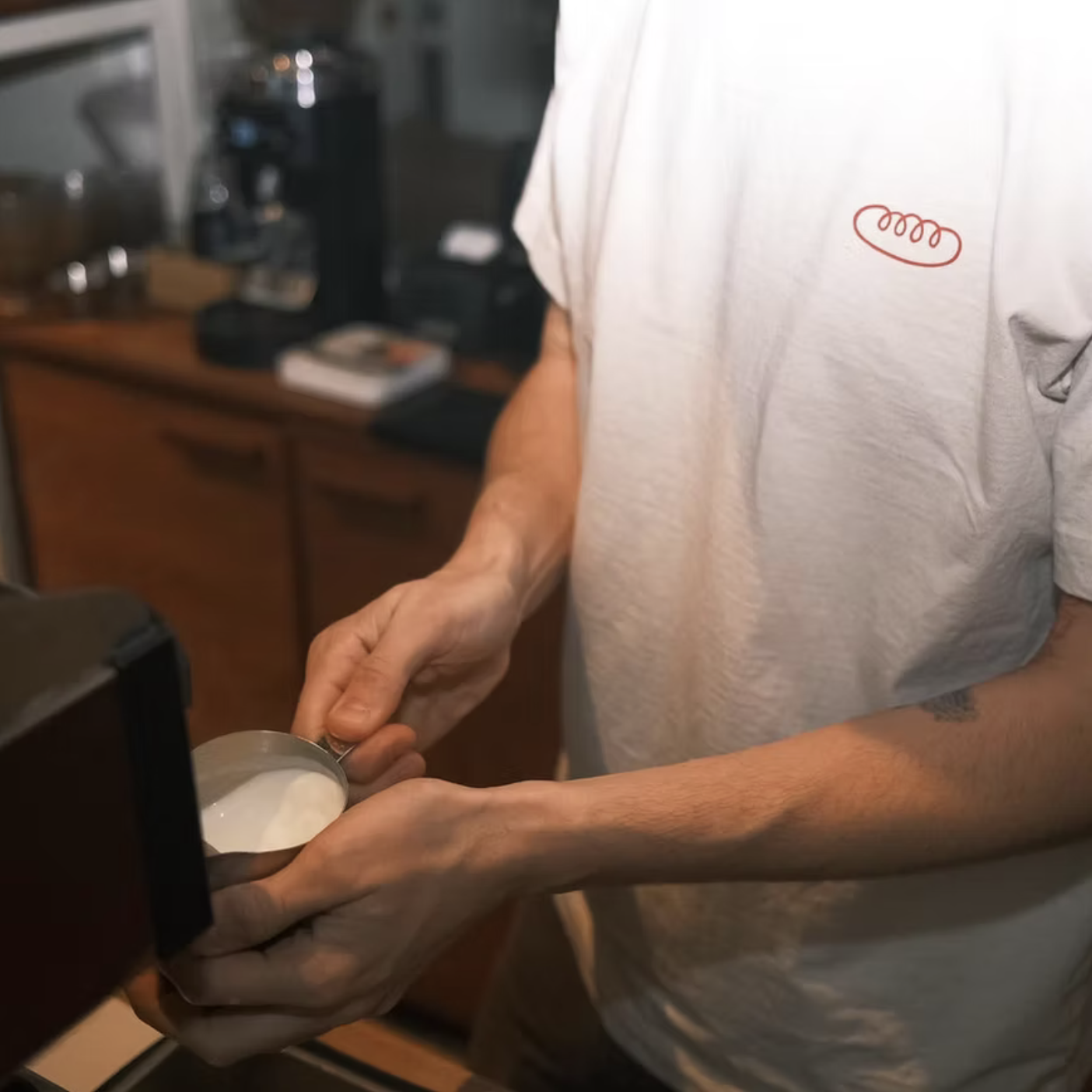 A person holding a metal pitcher of milk in a kitchen, wearing a white t-shirt with a small red embroidered logo.