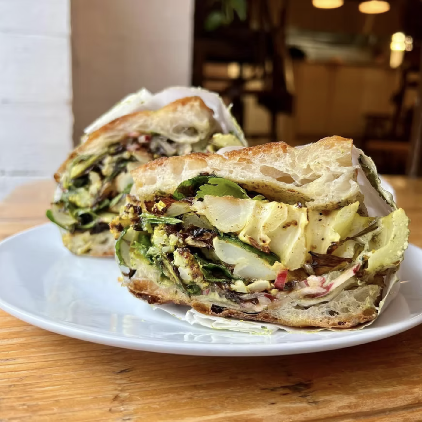 Cross-section of a vegetable-filled sandwich with leafy greens, cucumbers, and other vegetables on a white plate, on a wooden table in a restaurant setting.