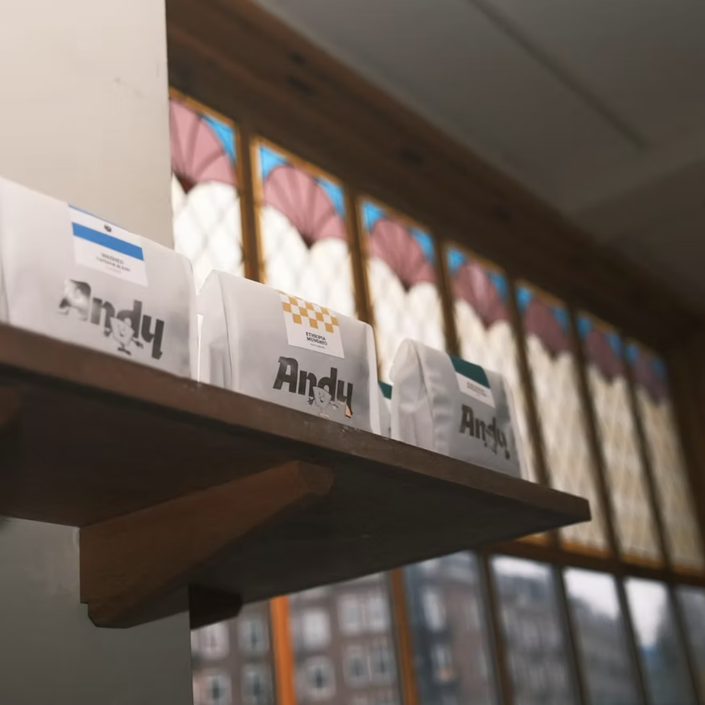 Shelf with white paper bags labeled 'Andy' with cartoon characters, in front of stained glass windows with pink and white design.
