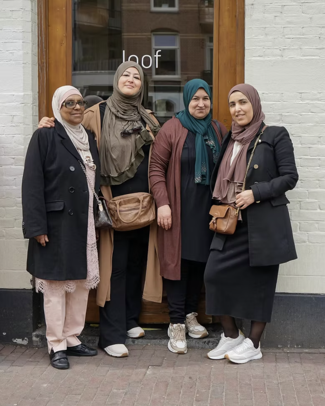 Four women standing in front of a white brick wall, posing for the photo. They are wearing modest clothing and headscarves.