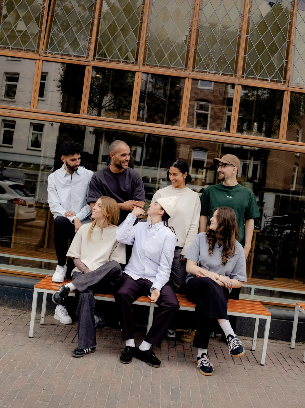 Group of seven diverse young adults sitting and standing on a bench outside a building with large windows, smiling and engaging in conversation.