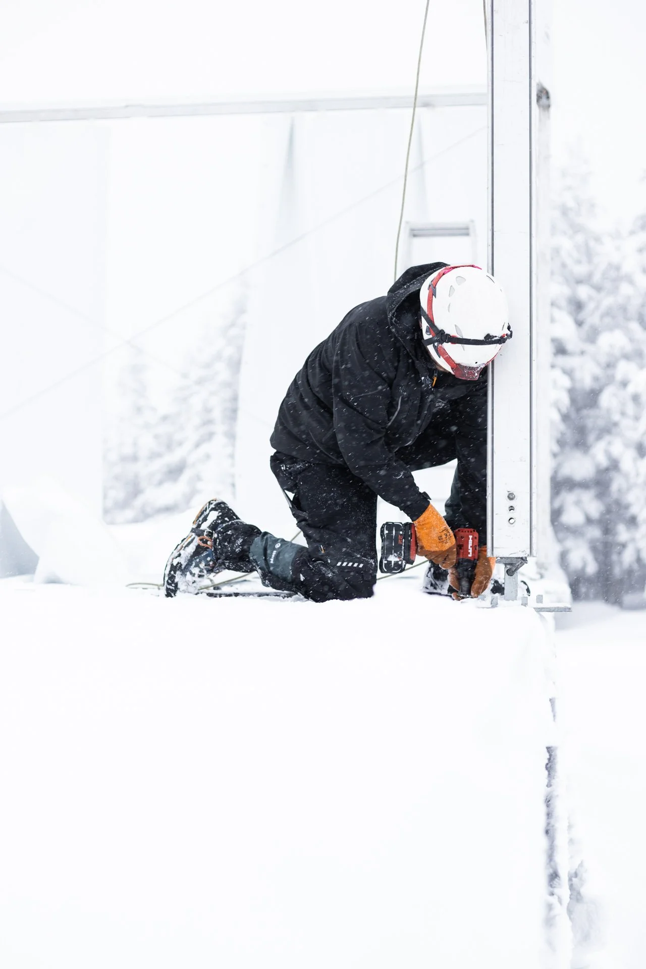 Ein Mann in Winterkleidung arbeitet mit Werkzeug im Schnee, trägt einen Schutzhelm und Handschuhe.