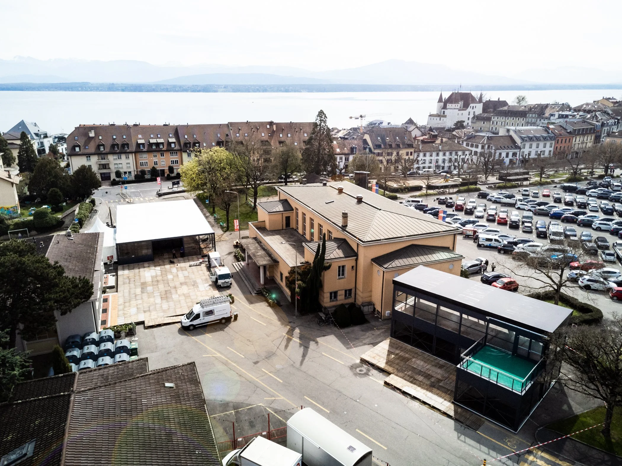 Luftaufnahme von einem Temporärbau mit Festfassaden, Parkplatz mit Autos, See im Hintergrund und Berge im Flachland.