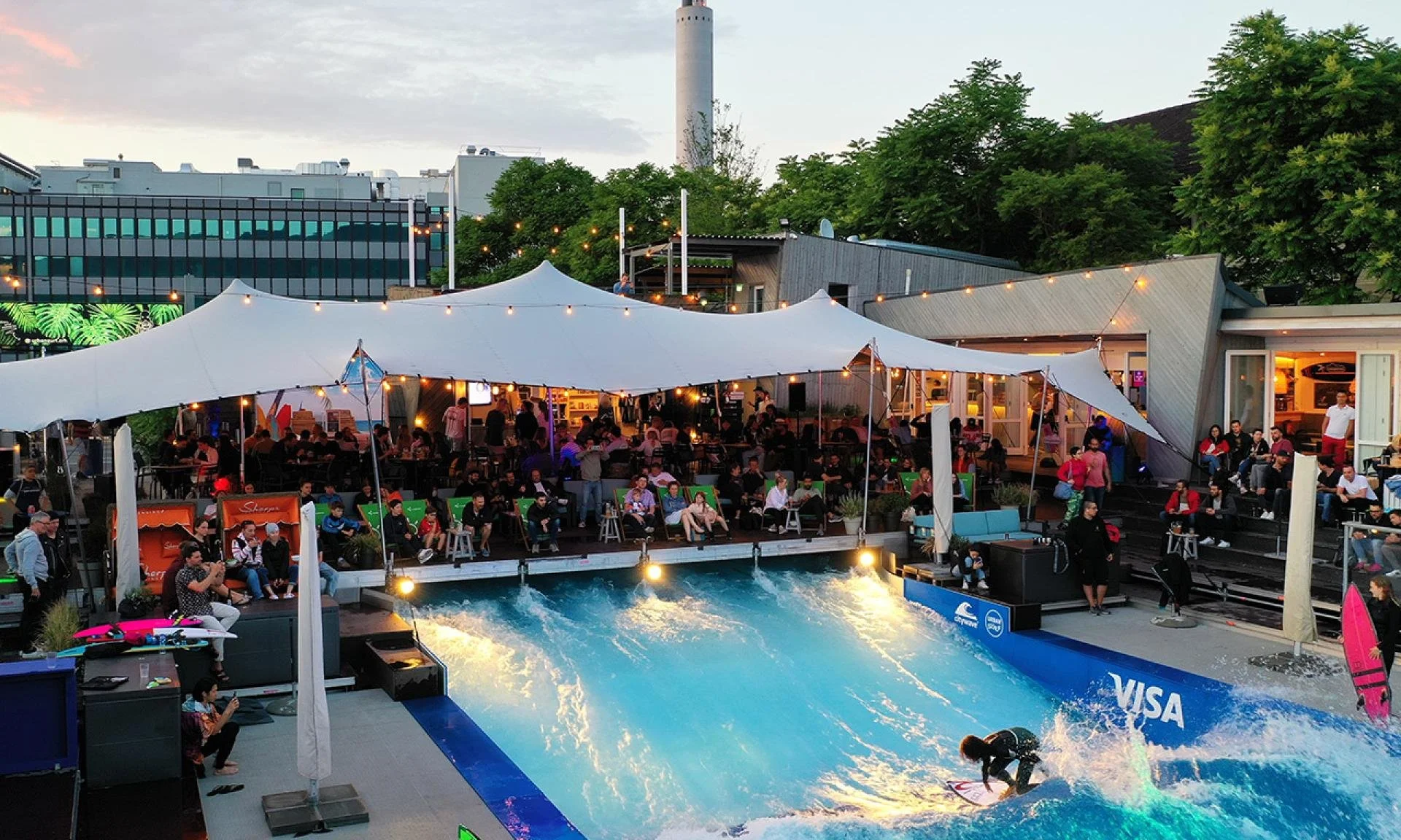 Menschen beim Surfen in einer künstlichen Welle in einem Outdoor-Unterhaltungskomplex bei Sonnenuntergang, umgeben von Sitzgelegenheiten und Zelten, mit grünen Bäumen und Gebäuden im Hintergrund.