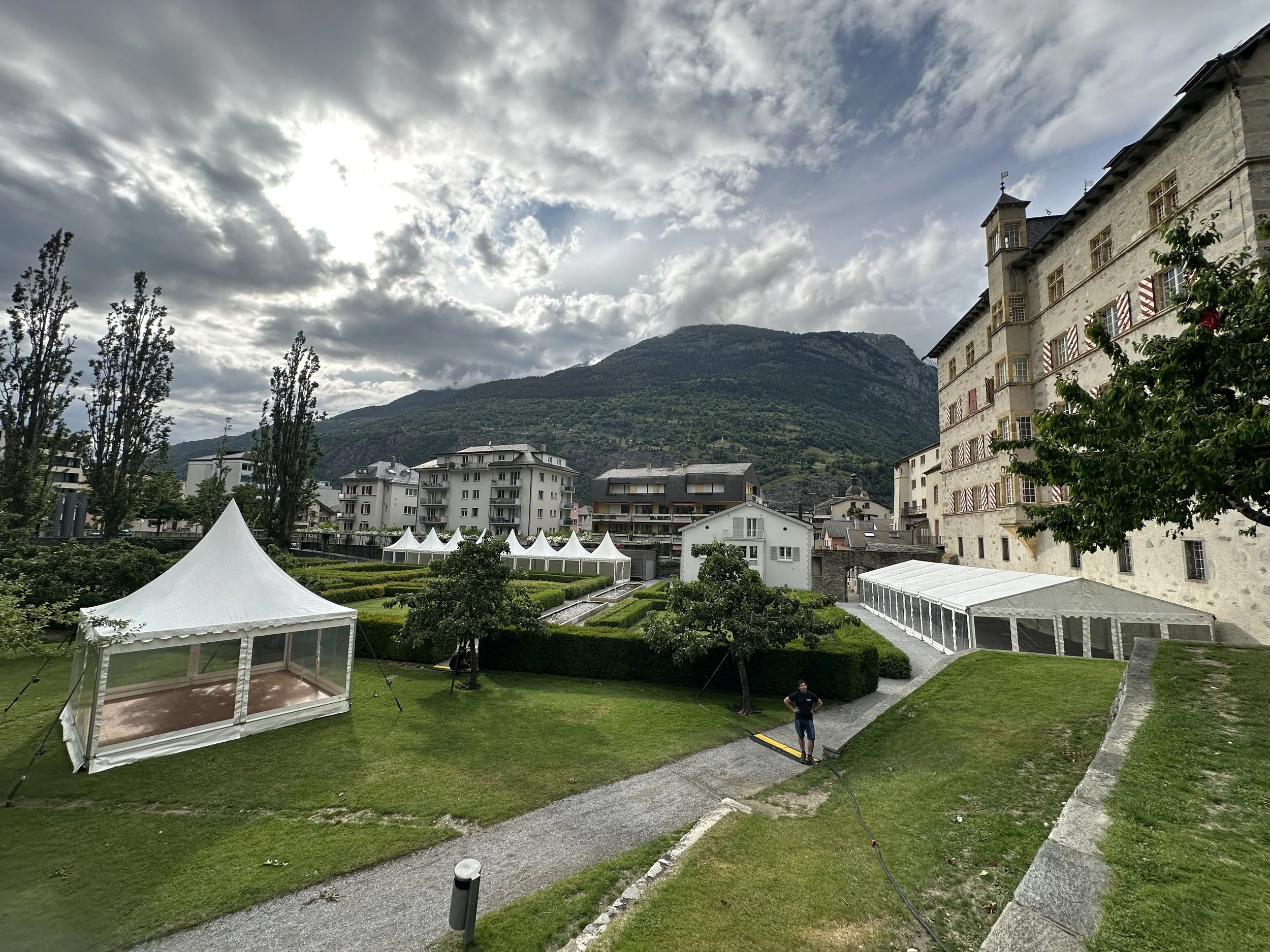Ein Garten mit weißen Zeltbauten, gepflegten Beeten und Bäumen vor historischen Gebäuden und Bergen im Hintergrund, bewölkter Himmel.