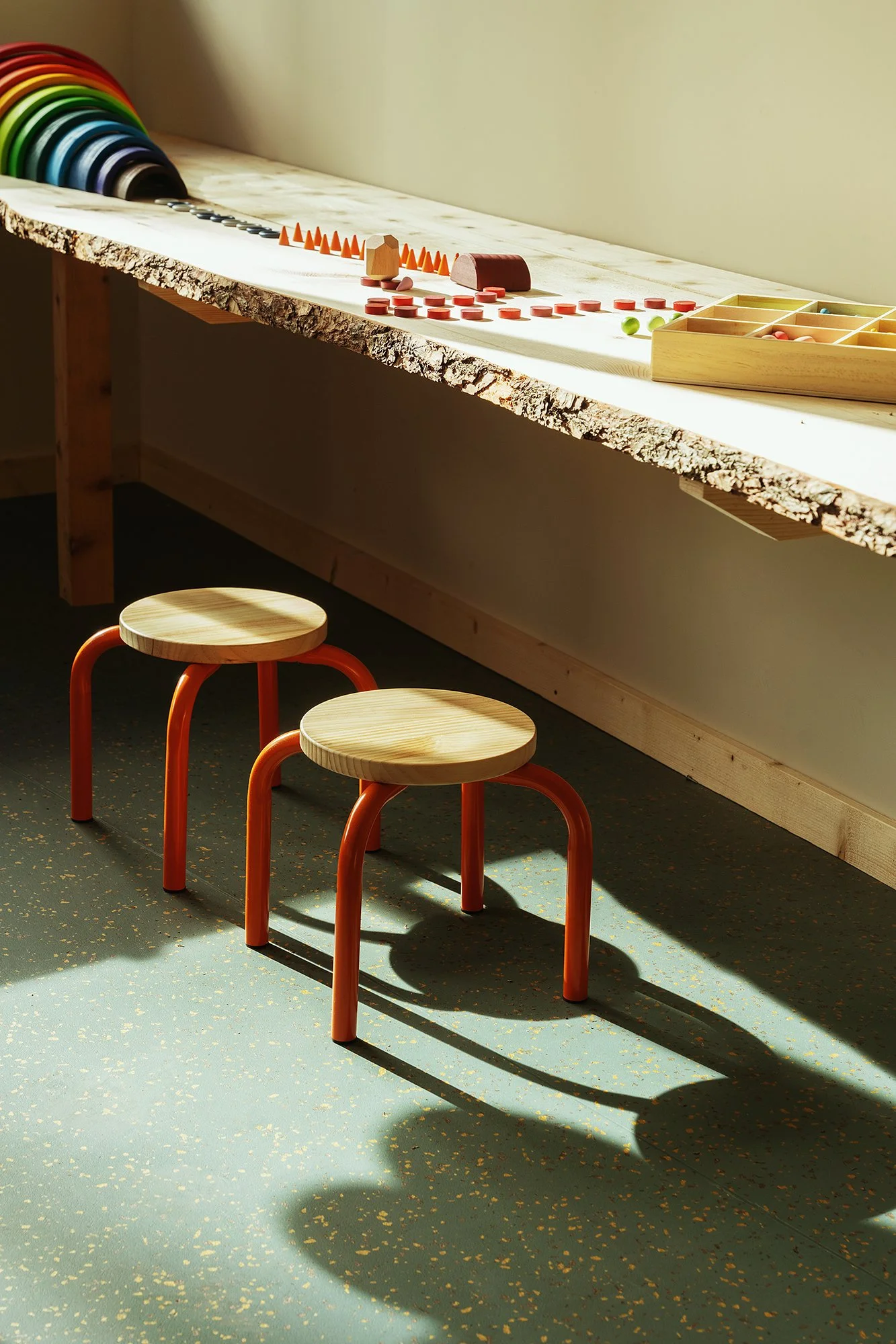 Children's play area with three wooden stools with orange legs, a worktable with various game pieces and rings, and a set of colorful curved rods, with sunlight streaming in.