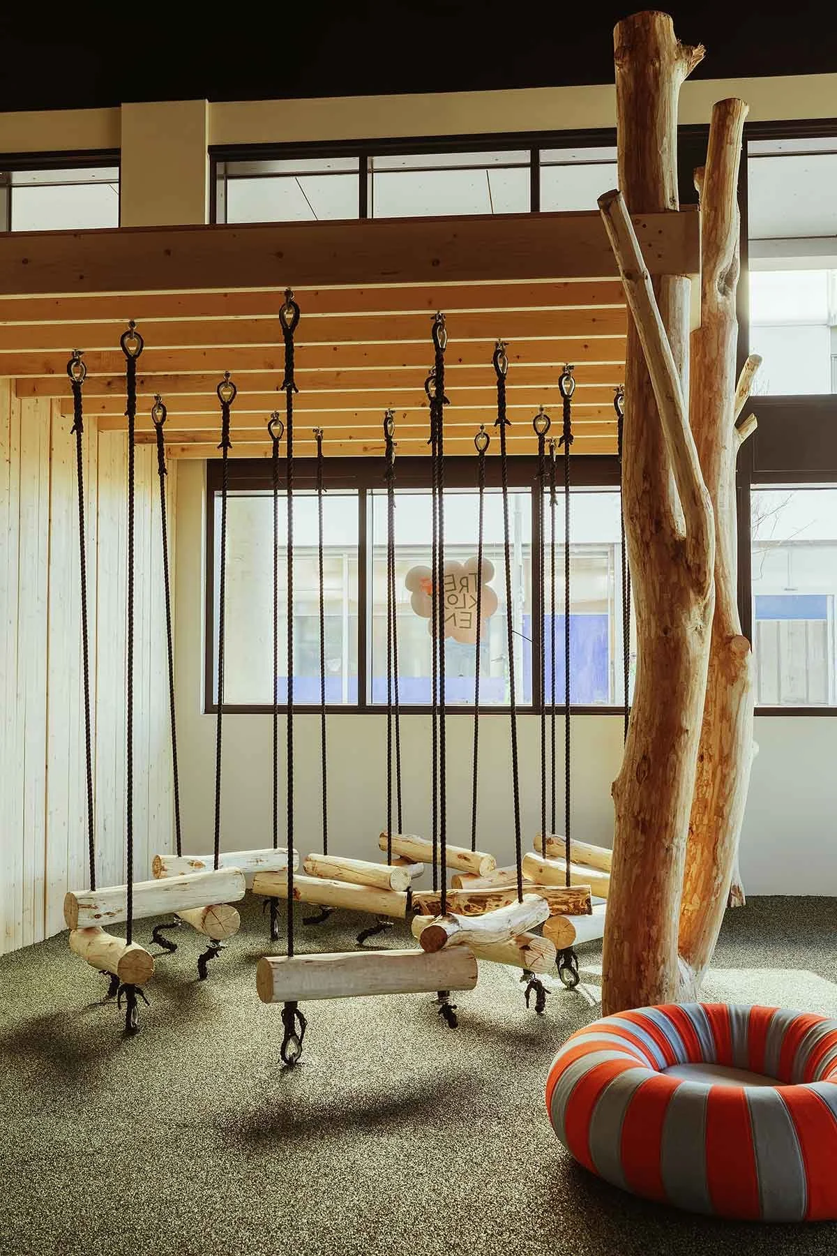 Indoor play area with wooden swing hanging by ropes and a tree trunk support, a colorful striped circular seat, and large windows with natural light.