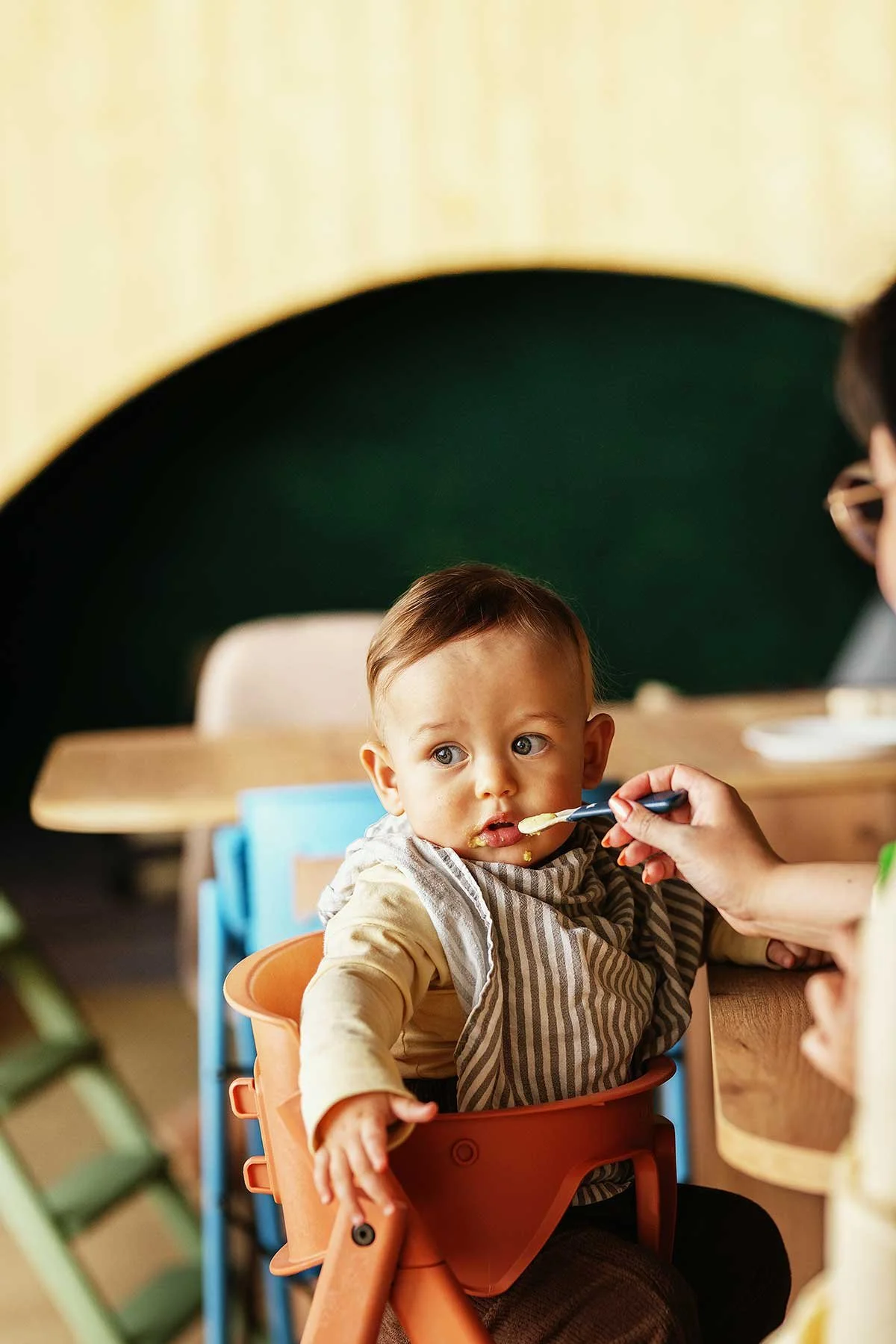 A young boy sitting in a high chair being fed by an adult, with food around his mouth, in a cozy dining area with a rounded green wall and yellow ceiling.