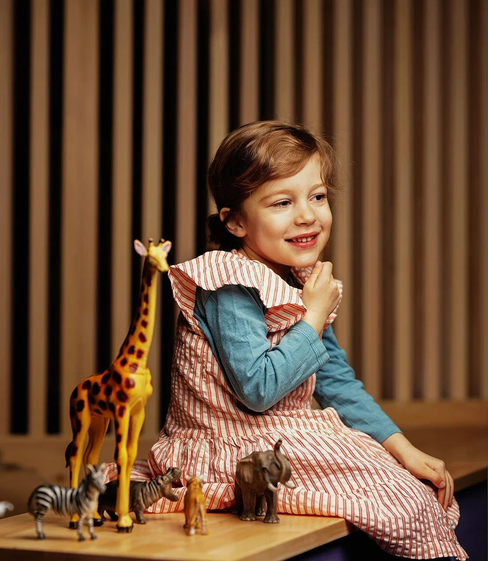 A young girl sitting on a wooden surface, smiling with her head slightly tilted and hand touching her chin. She is surrounded by toy animals including a large giraffe, zebra, elephant, and lion. The background features vertical wooden slats.