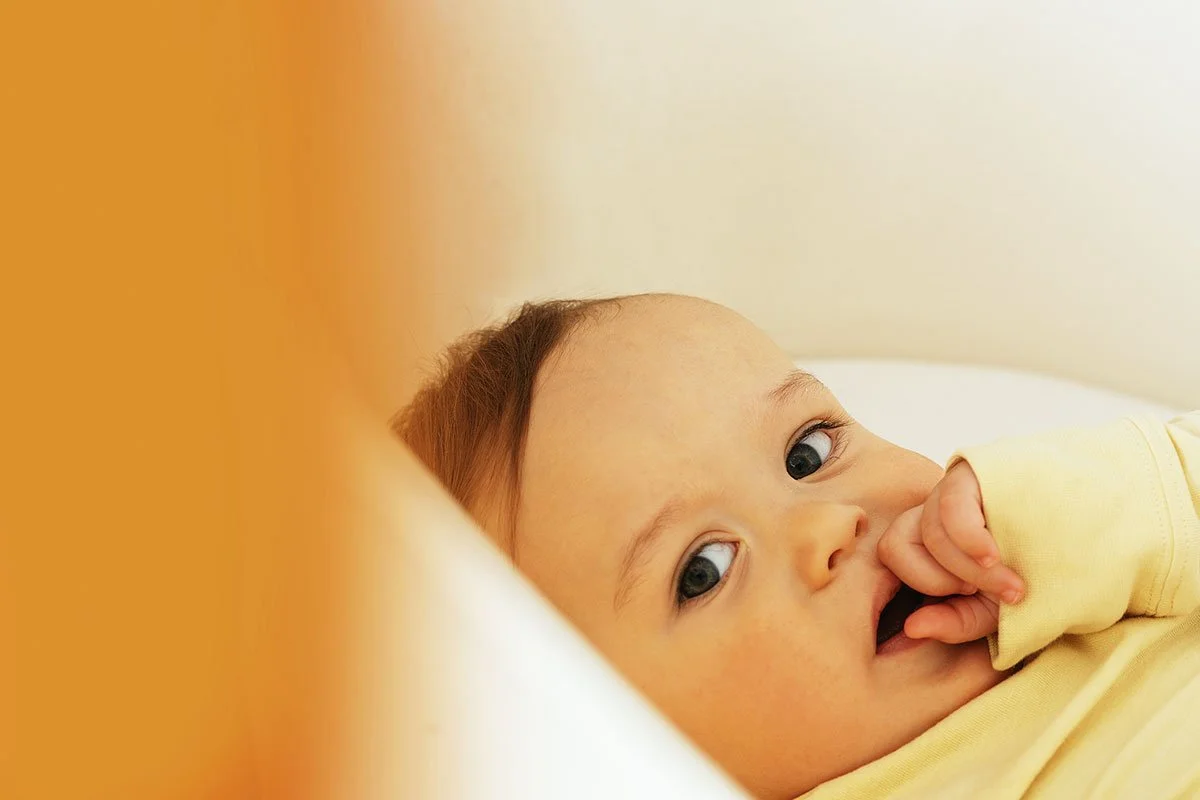 A close-up of a young baby lying in bed with a hand in their mouth, looking at the camera with blue eyes, wearing a yellow outfit.