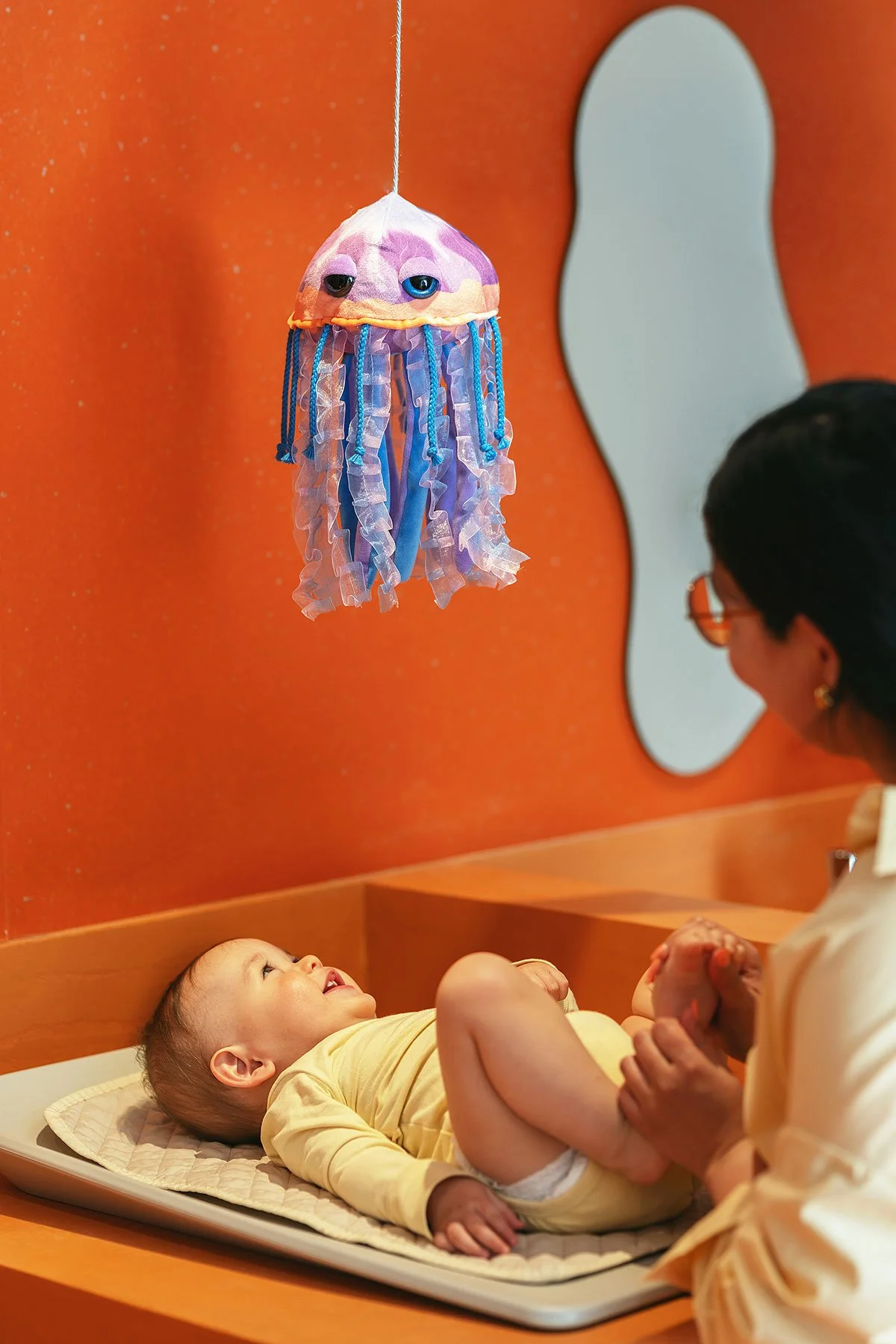 A smiling baby lying on a bed looking at a hanging plush jellyfish toy while an adult woman, possibly the baby's mother, interacts with the child in a room with orange walls and a mirror.
