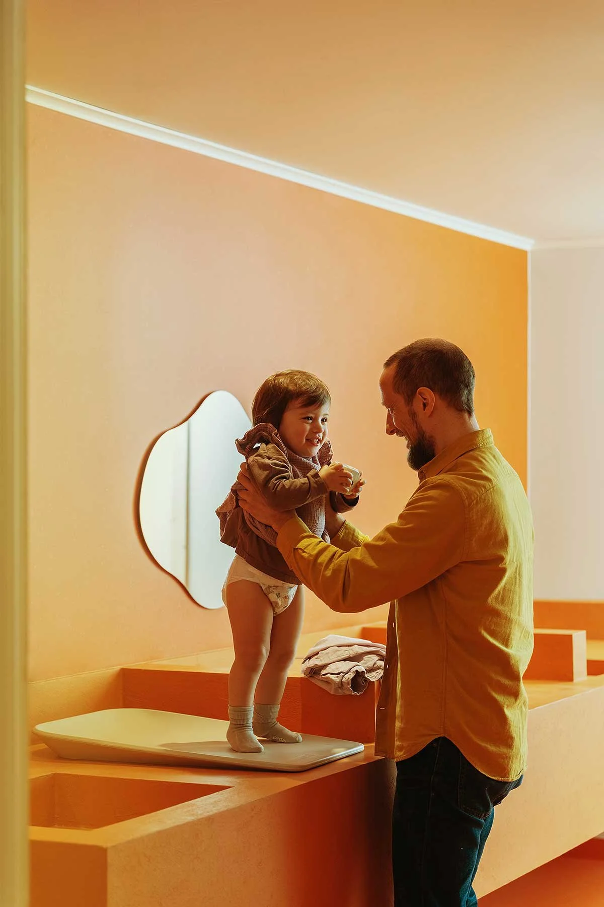 A father helps his young daughter stand on a scale in a warm-colored room. The girl is smiling and holding a cup, wearing a brown jacket and diapers. There is a mirror on the wall behind her, and folded clothes on the surface next to the scale.