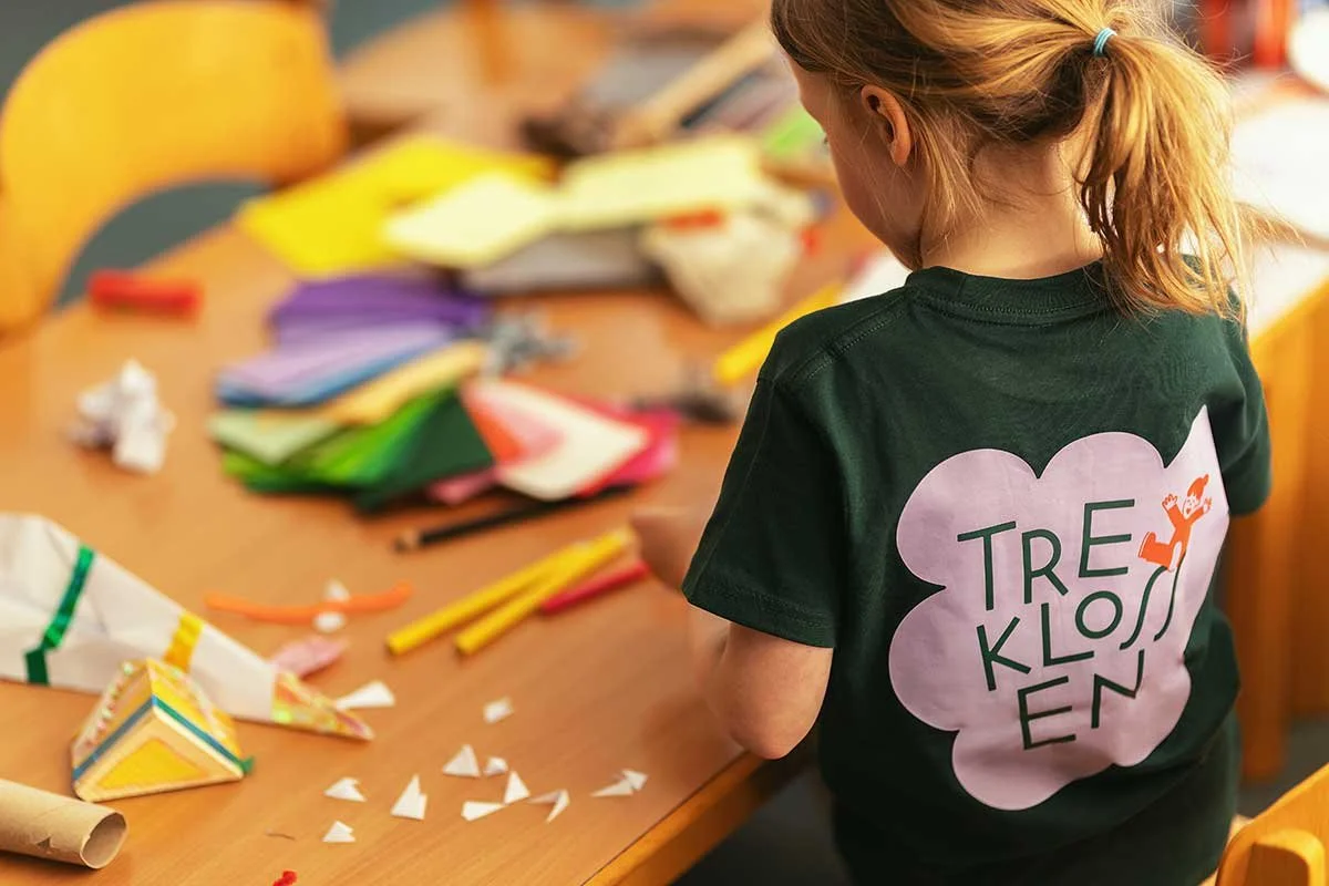 A young girl with red hair tied in a ponytail, wearing a green Trekklossen shirt, standing at a table covered with colorful craft supplies, paper, and scissors, working on a craft project.