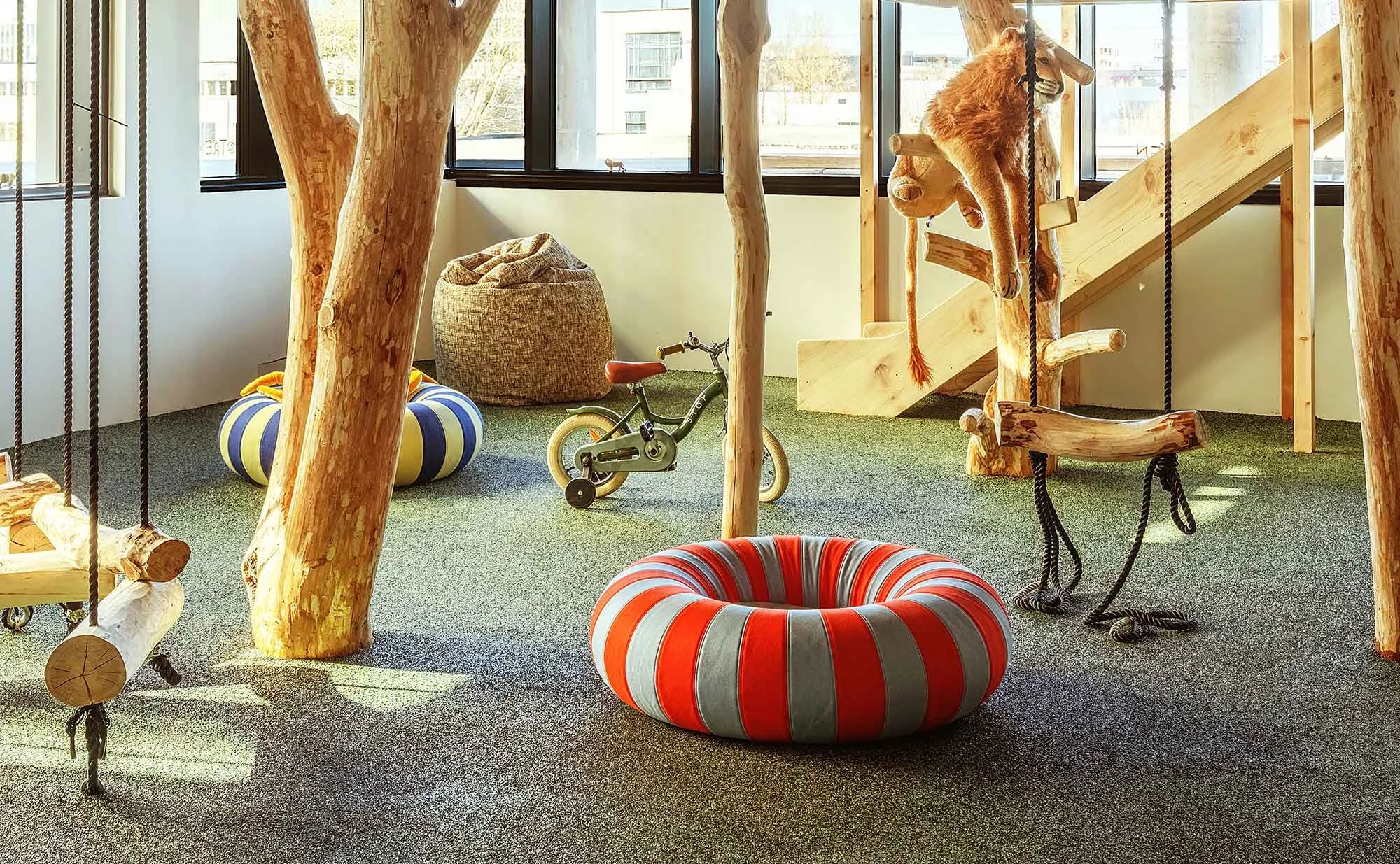 Indoor playroom with wooden structures, swings, a small bicycle, and colorful cushioned seating, illuminated by large windows.