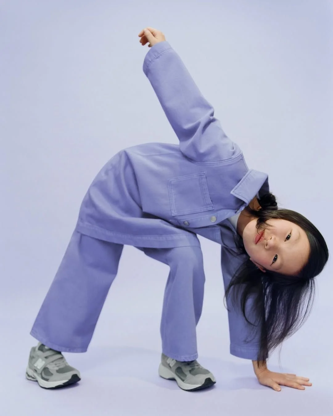 Young girl in oversized purple jacket and pants, wearing sneakers, posing in a backward bend with one hand on the ground and the other raised, against a plain light background.