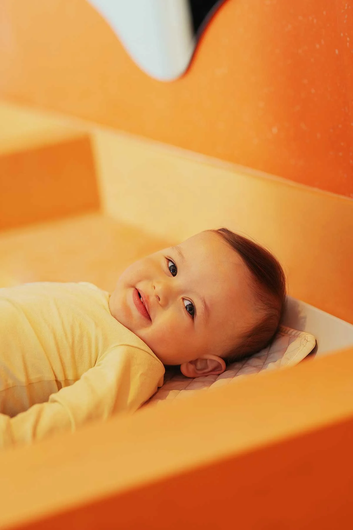 A young child lying in bed, smiling and looking at the camera with a happy expression, surrounded by orange bedding and wall.