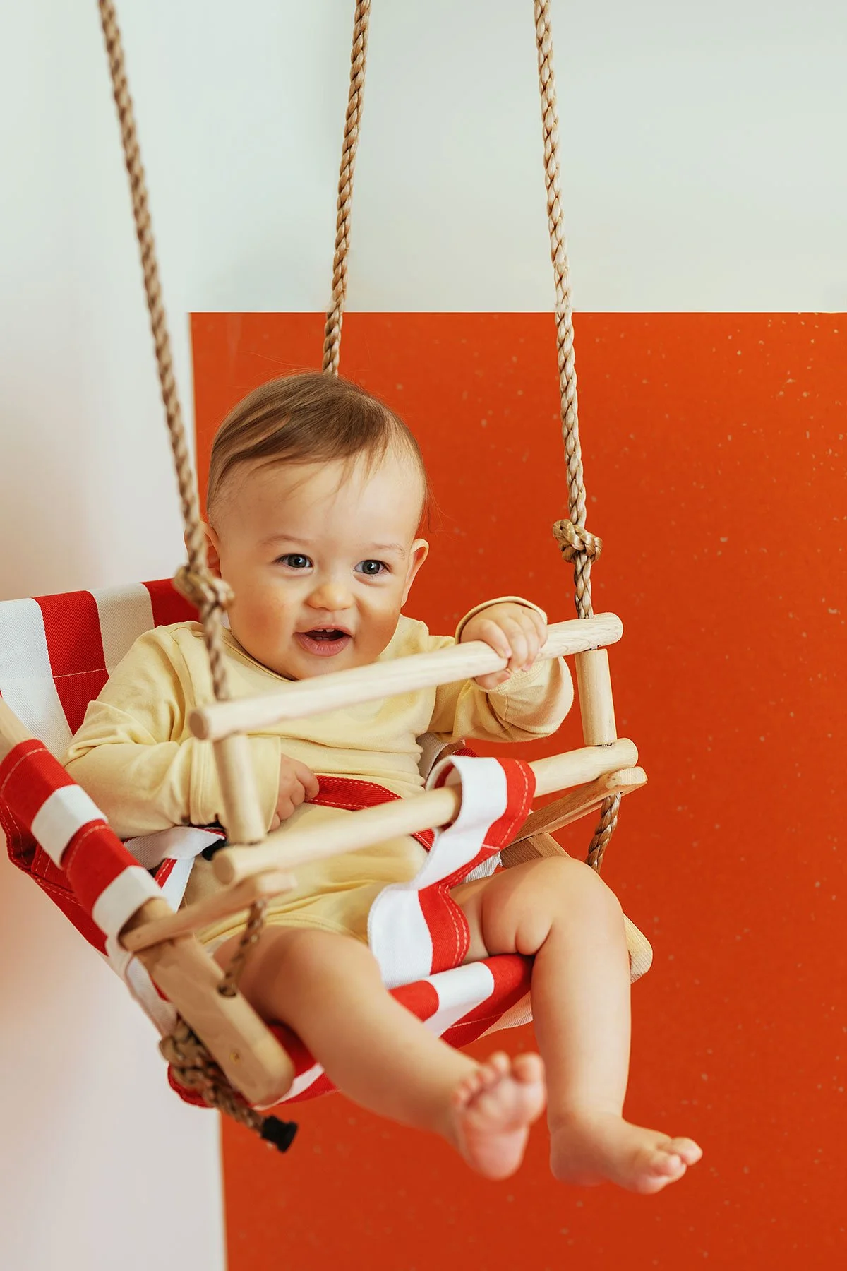 A smiling baby sitting in a red and white striped swing with beige ropes in front of an orange and white wall.