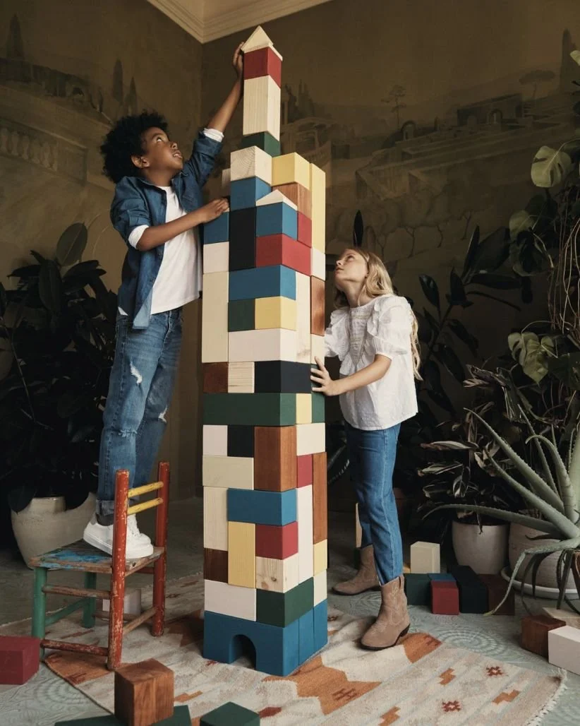 Two children, a boy and a girl, building a tall tower with colorful wooden blocks indoors, with large plants and a decorative background.
