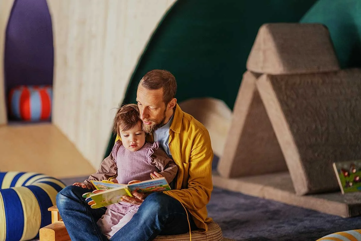 A man reading a children's book to a young girl sitting on his lap in a colorful indoor play area.