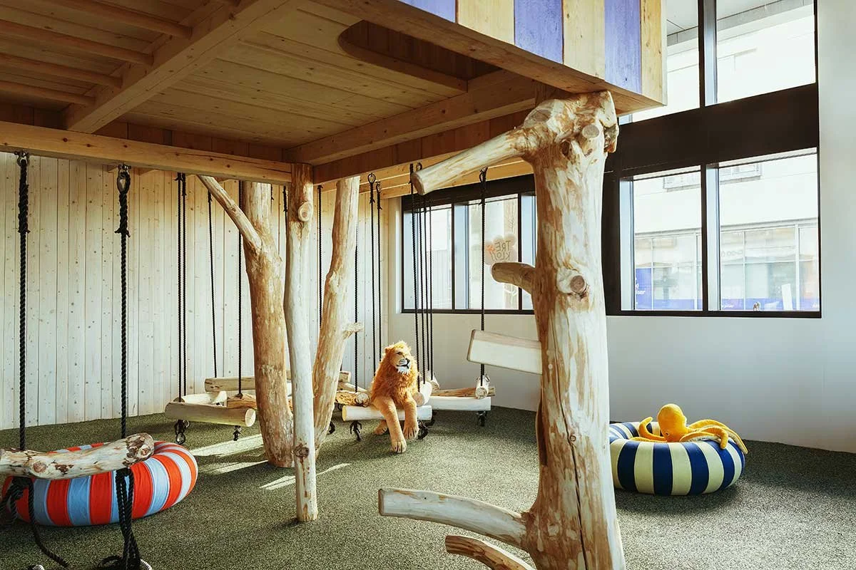 Indoor play area with wooden tree-like structures, swings, and cozy pet beds. A stuffed lion toy is sitting on a swing.