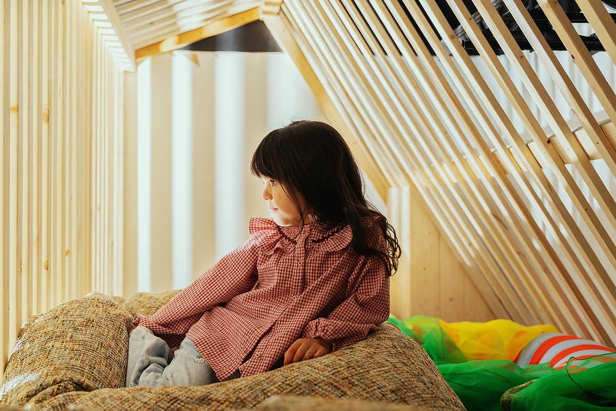 A young girl with black hair, wearing a red checkered dress, sitting on a multicolored bed inside an attic with wooden framing and slanted ceiling.