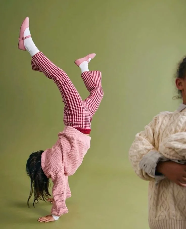 Child doing a handstand with legs raised in the air, wearing pink and red checkered pants, a pink sweater, and pink shoes with ankle straps, against a green background. Another person partially visible, wearing a beige sweater, is standing nearby.