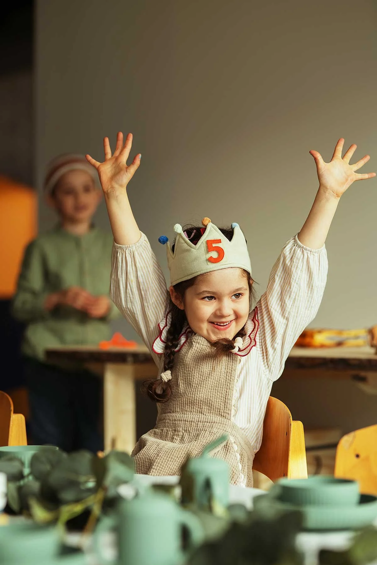 A young girl wearing a birthday hat with a red number 5 and colorful pom-poms, smiling with her hands raised, at a celebration or party with guests and tableware in the background.